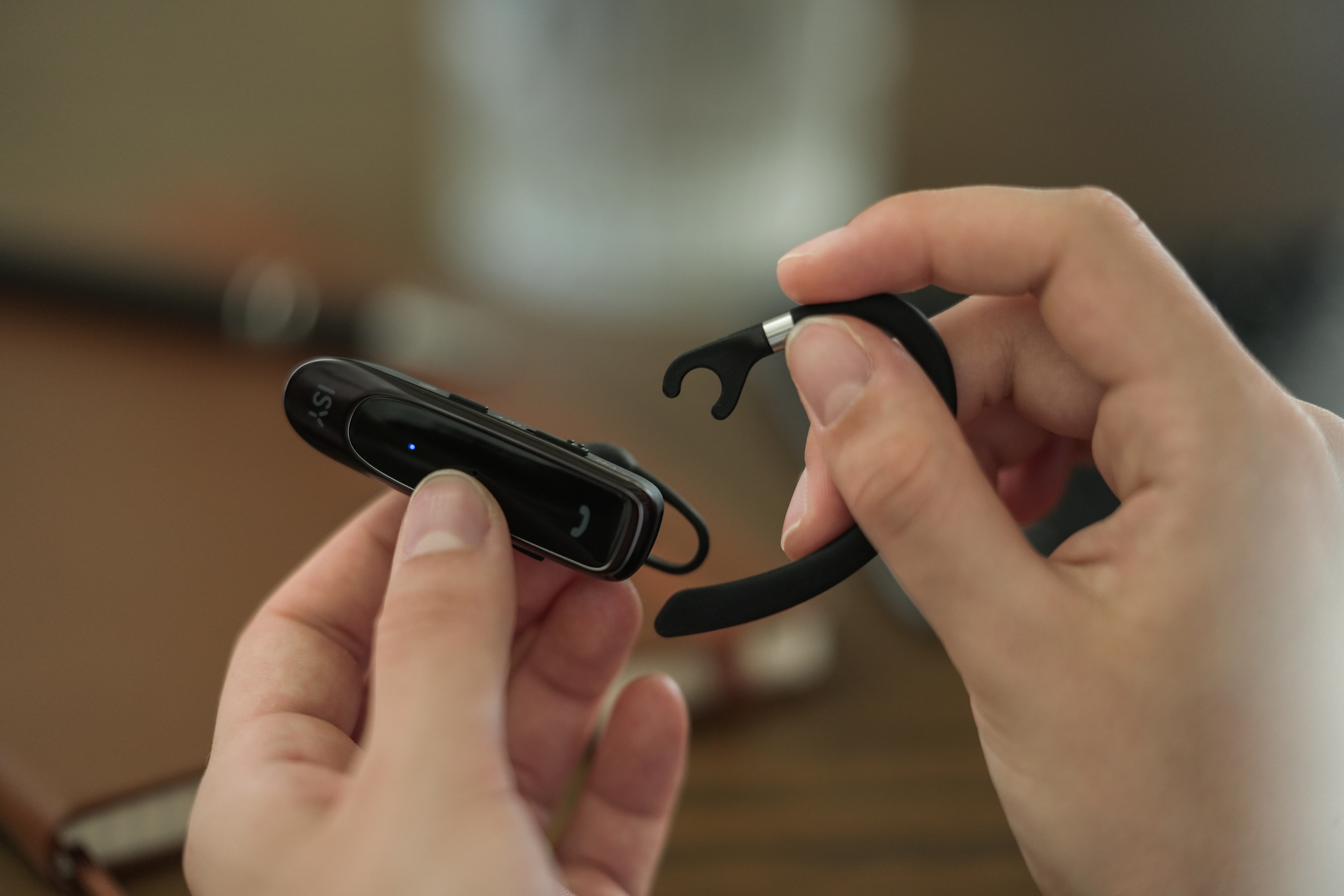 Hände halten ein schwarzes ISY Bluetooth-Headset. Das Headset wird zusammengebaut. Brauner Tisch im Hintergrund.