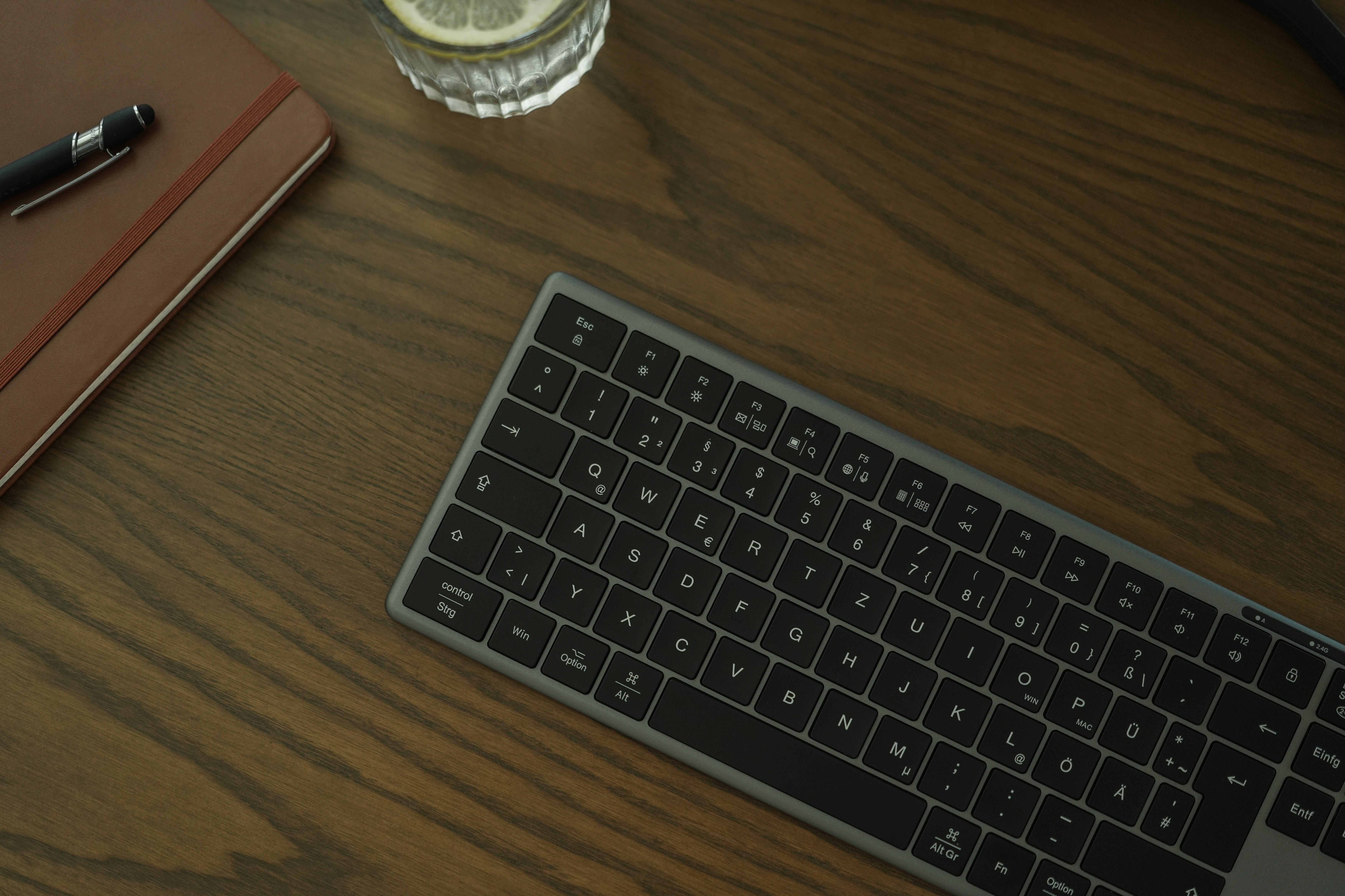 Eine Tastatur, ein Stift, ein Notizbuch und ein Glas Wasser mit Zitrone auf einem Holztisch.