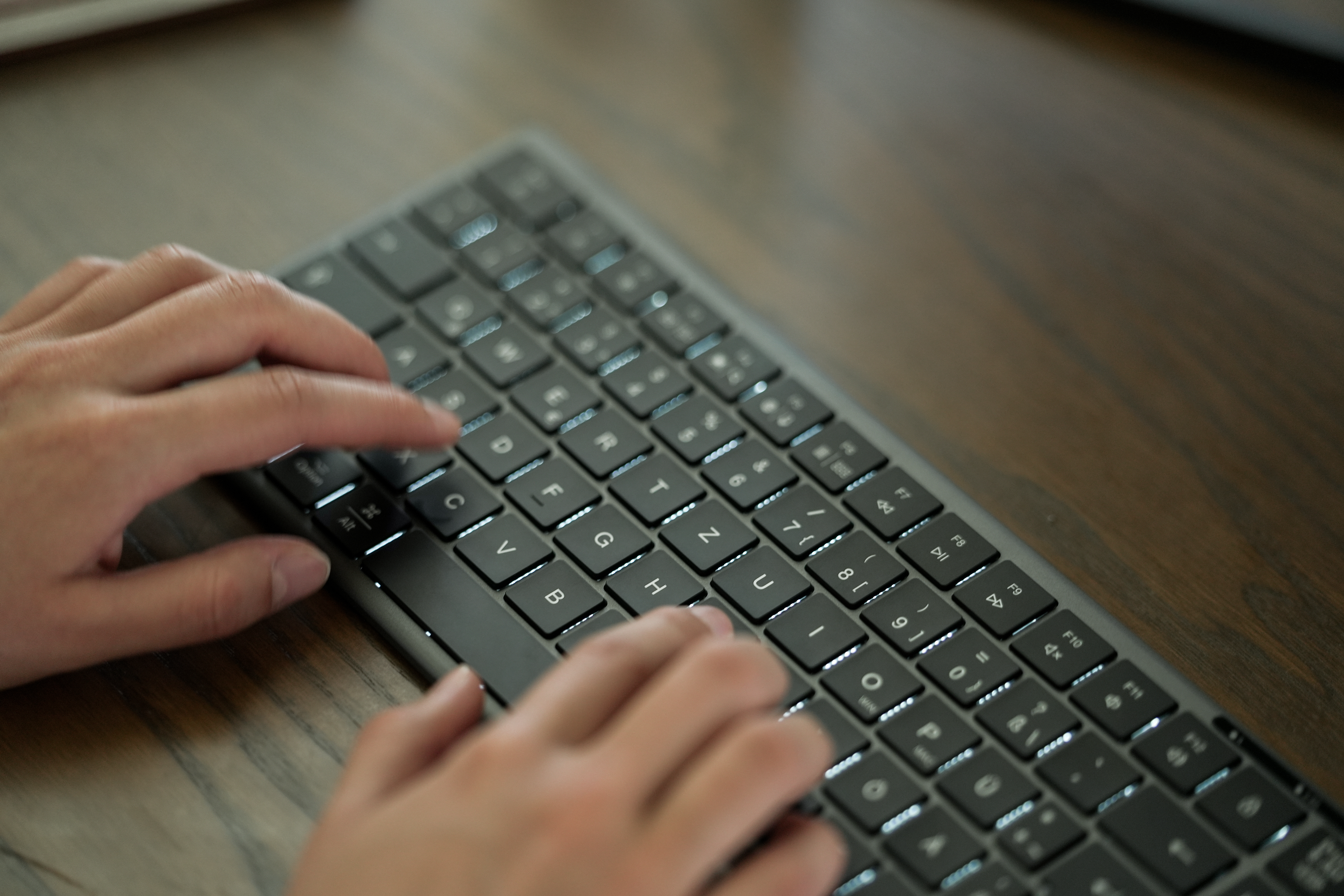 Hände tippen auf einer schwarzen Tastatur auf einem Holzschreibtisch. Tasten sind beleuchtet. Fokus liegt auf den Händen.