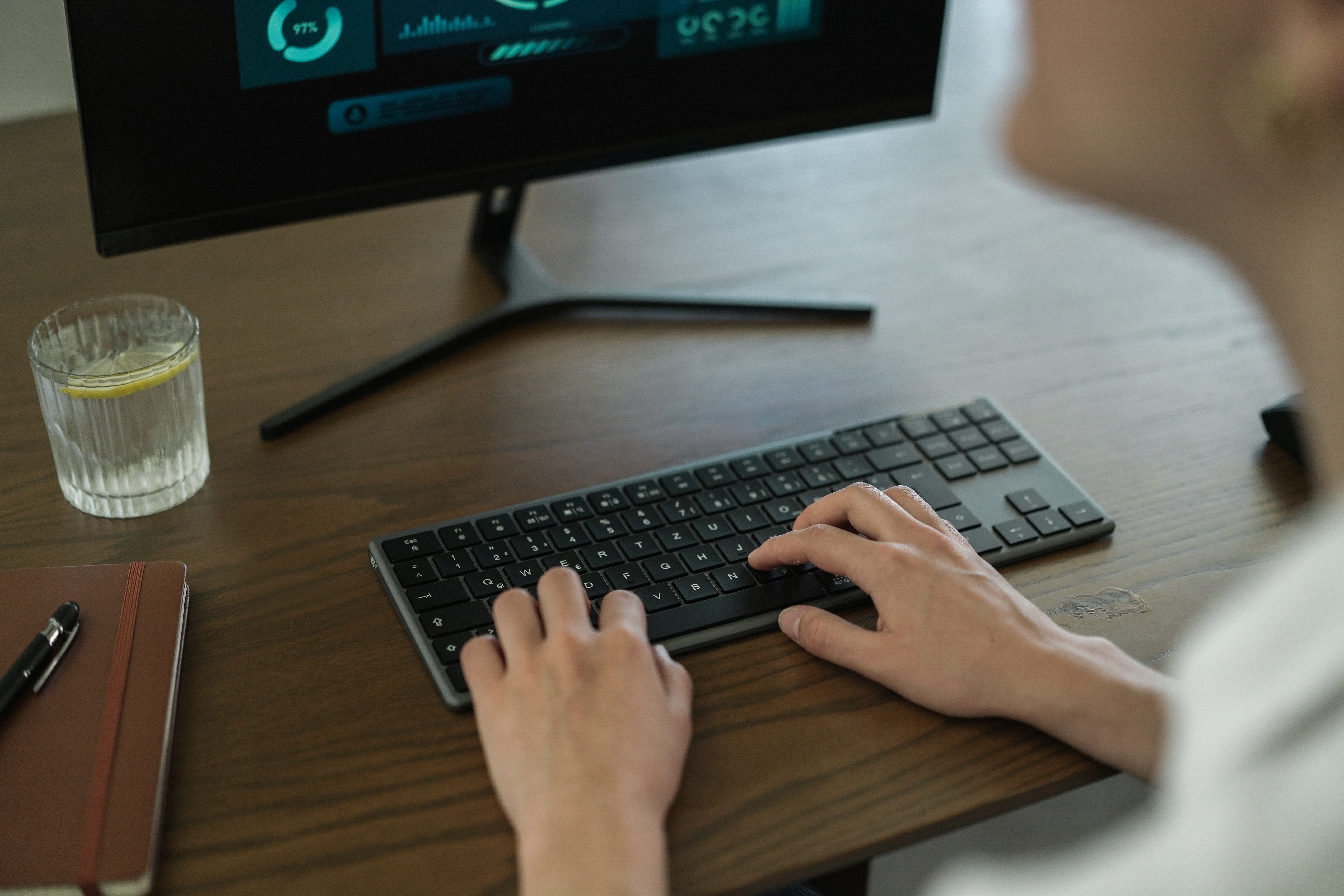 Person tippt auf einer Tastatur am Schreibtisch. Ein Monitor zeigt Daten. Glas Wasser mit Zitrone und Notizbuch.