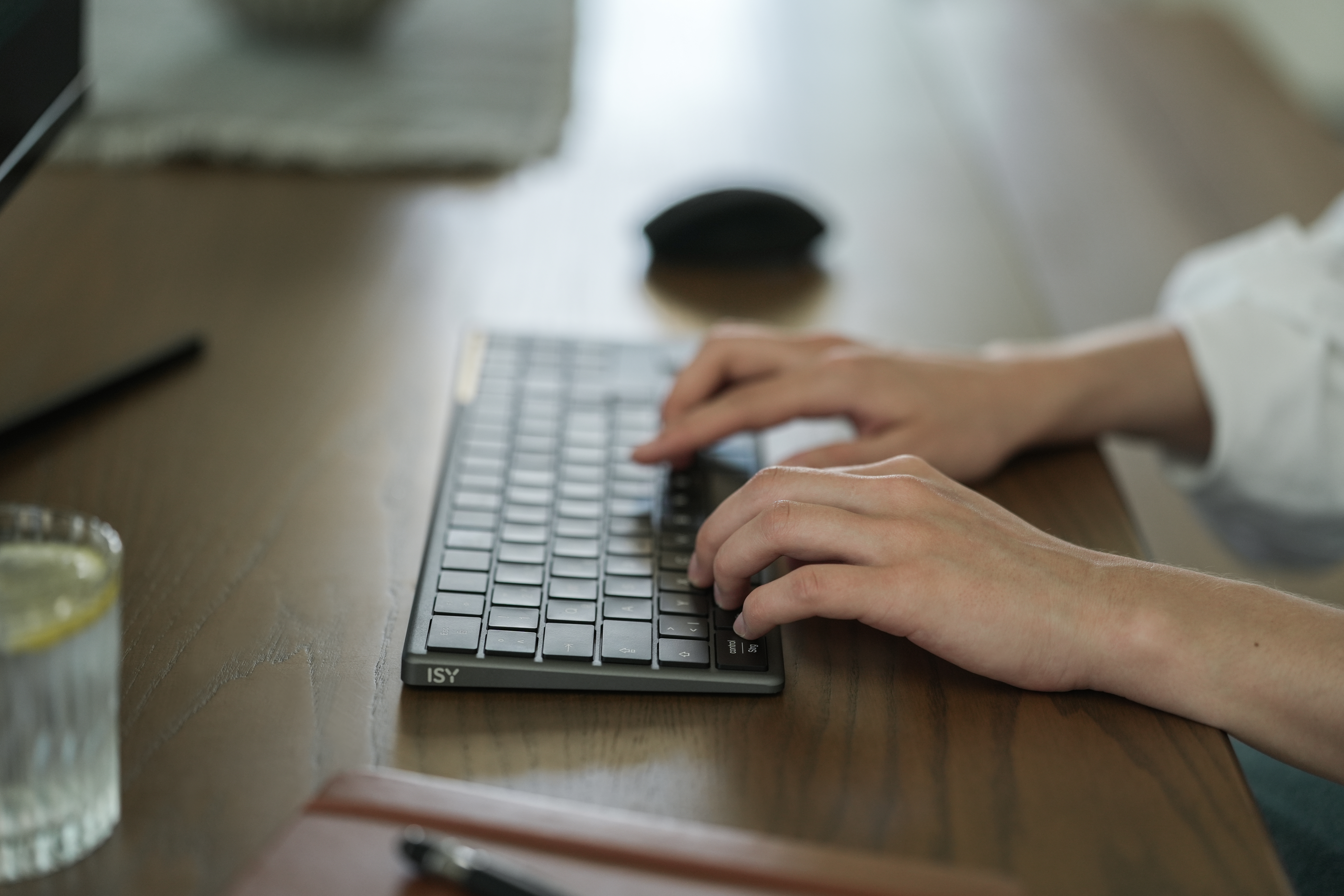 Hände tippen auf einer grauen Tastatur. Ein Glas mit Zitrone, eine Maus und ein Notizbuch liegen auf einem Holztisch.