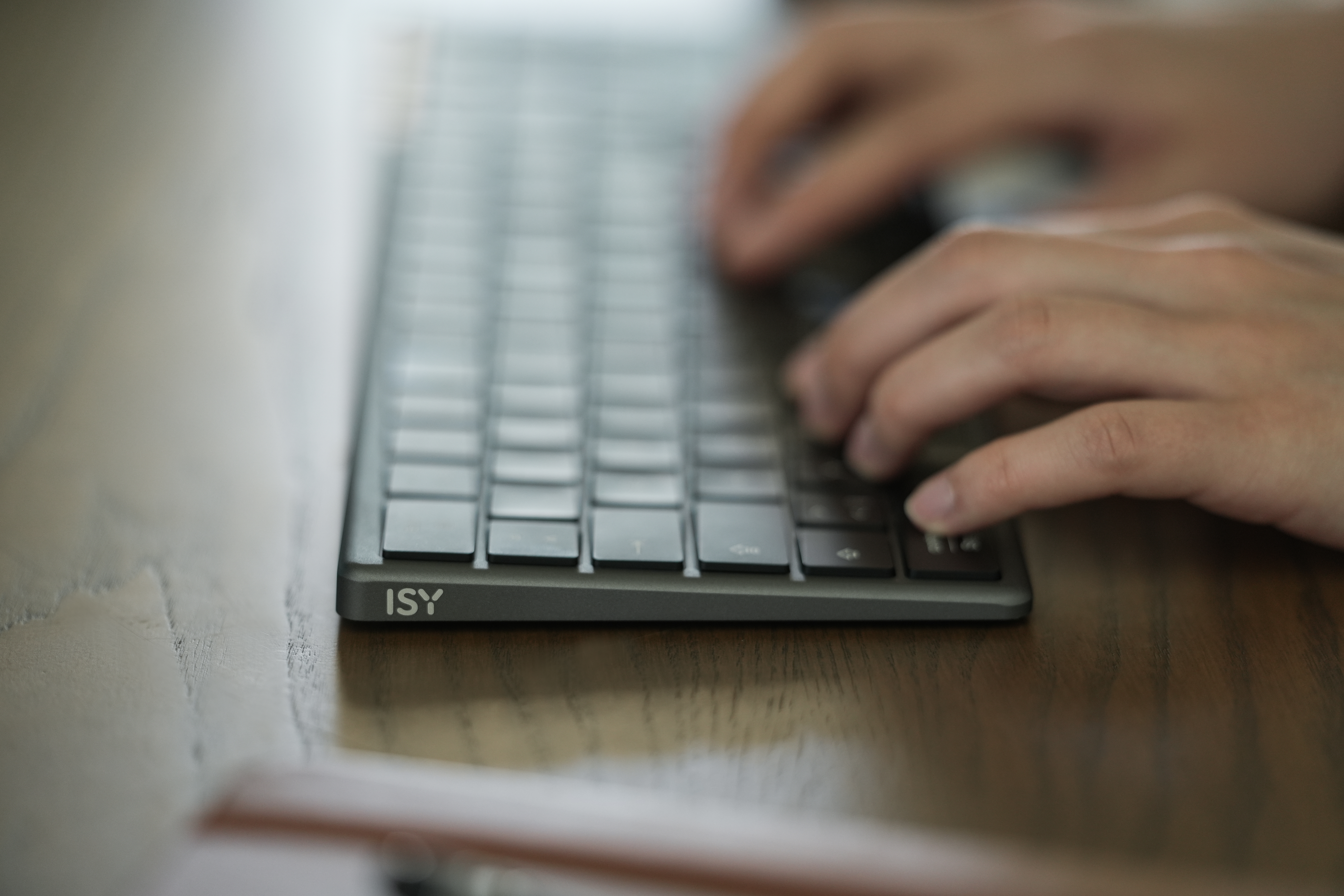 Hände tippen auf einer grauen ISY-Tastatur. Die Tastatur liegt auf einer Holzoberfläche. Der Hintergrund ist unscharf.