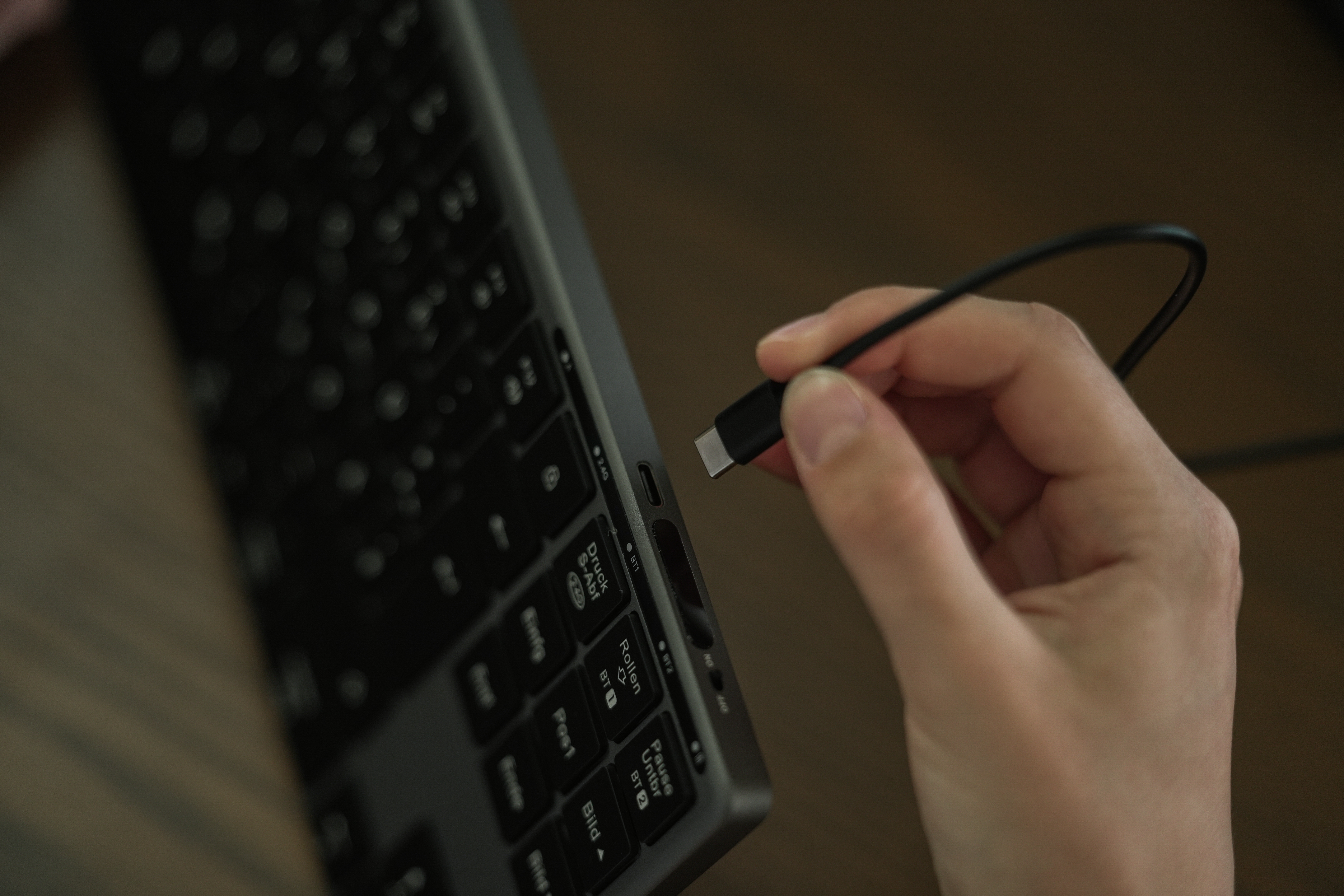 Eine Hand steckt ein USB-C-Kabel in eine Tastatur. Die Tastatur ist schwarz, der Hintergrund ist verschwommen braun.