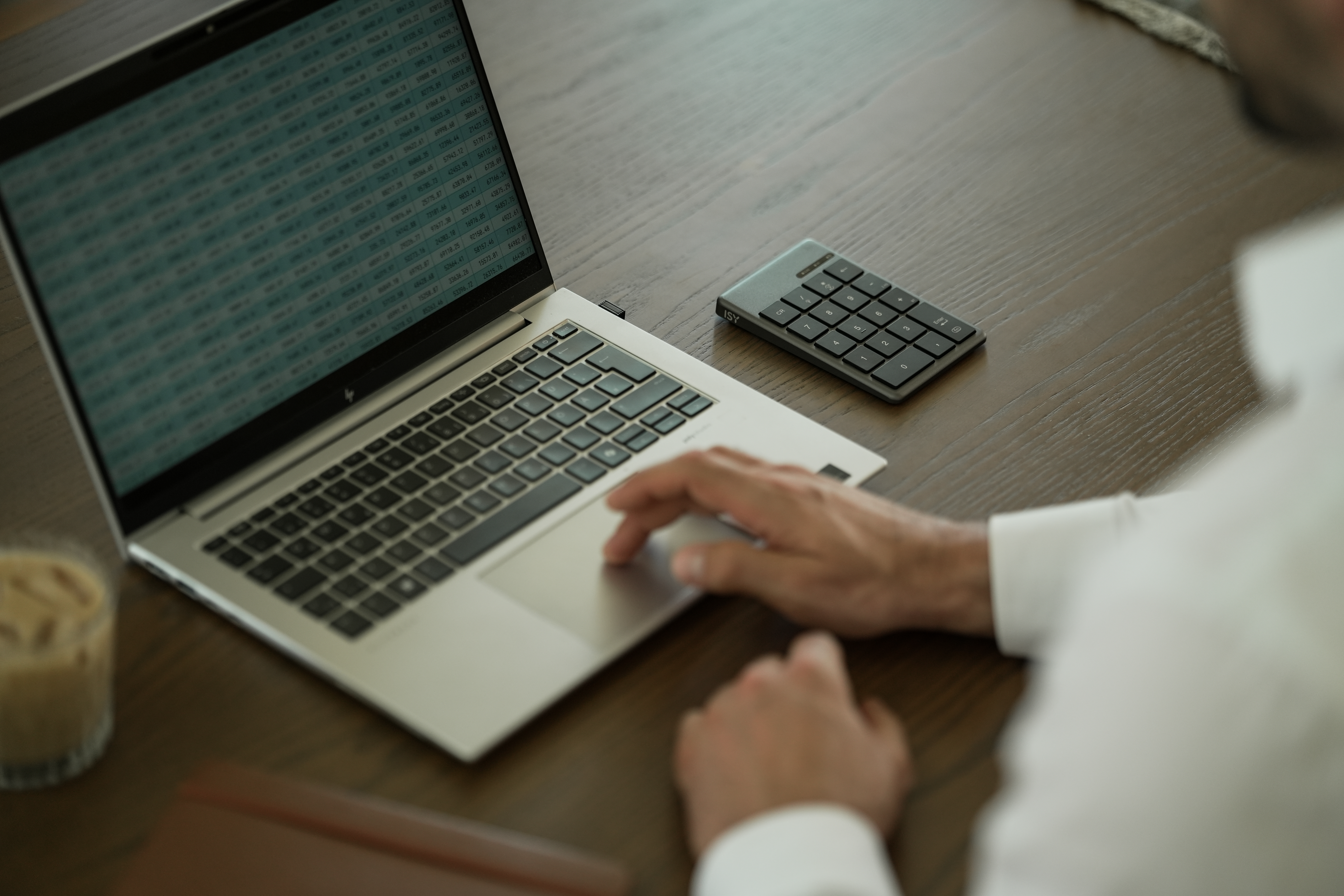 Ein Laptop zeigt Daten, ein Taschenrechner und ein Getränk auf einem Holztisch. Die Hand einer Person liegt auf dem Touchpad.