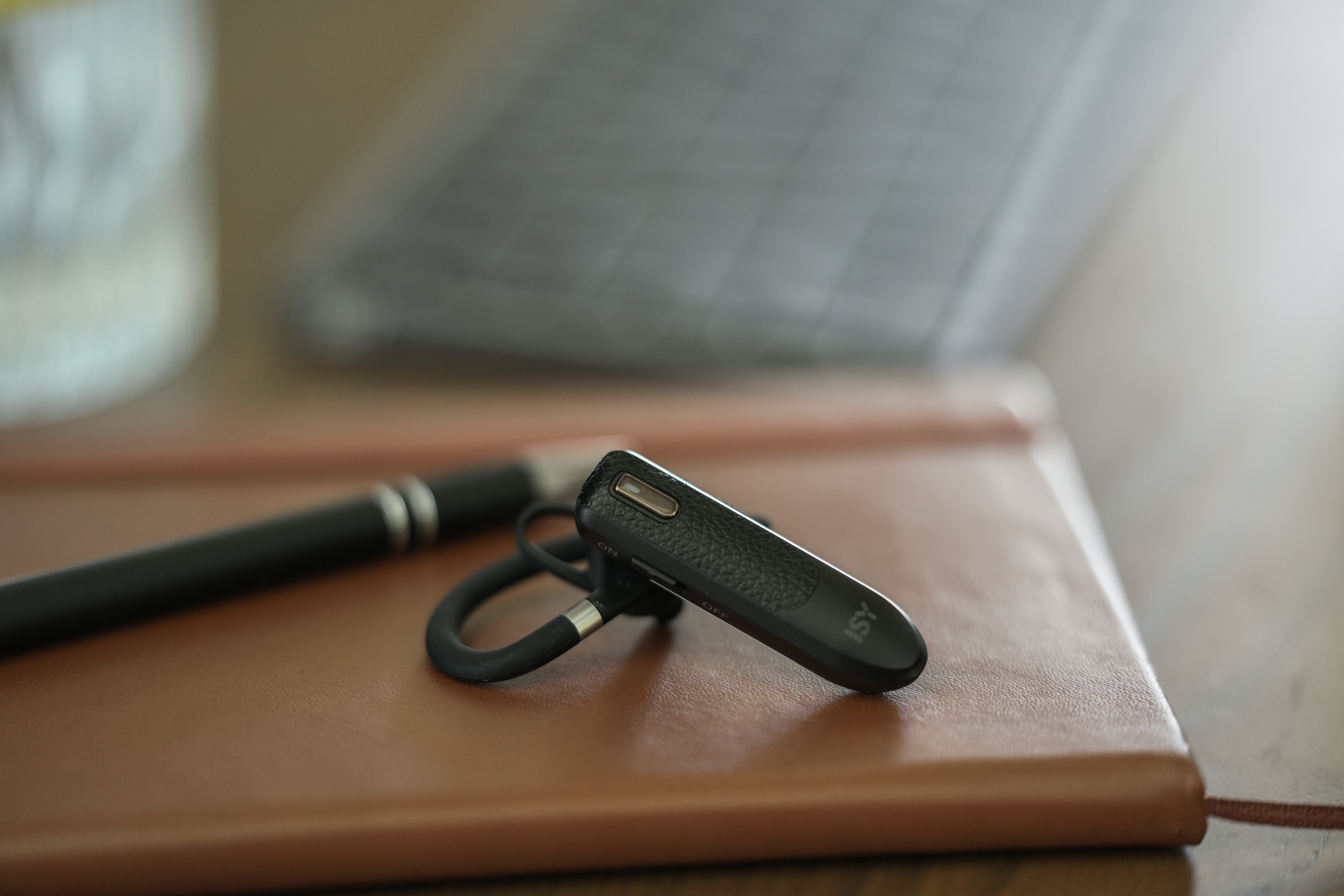 Ein schwarzes Bluetooth-Headset liegt auf einem braunen Notizbuch neben einem Stift. Im Hintergrund ist ein Laptop zu sehen.