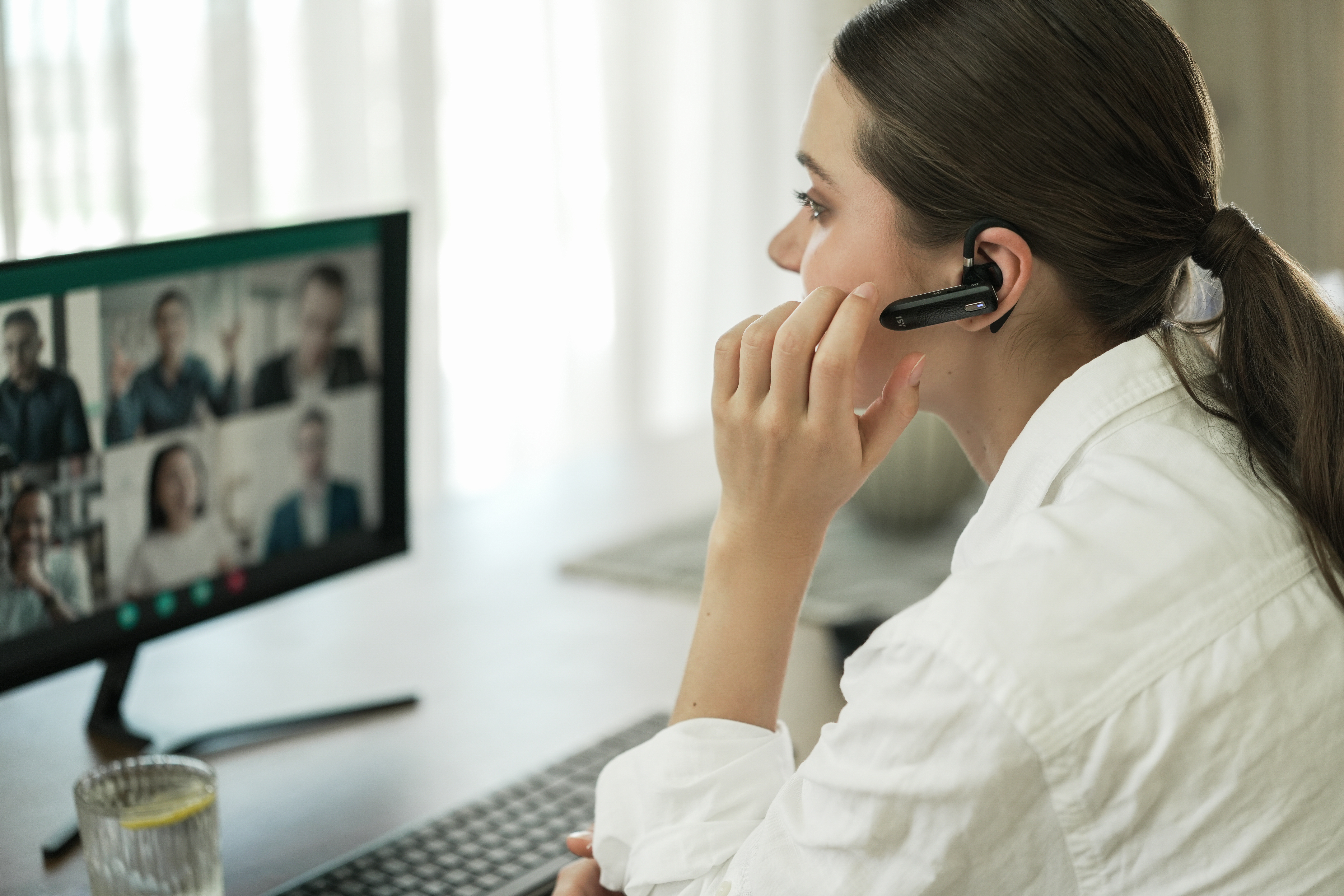 Eine Frau mit Headset nimmt an einem Videoanruf teil. Sie trägt ein weißes Hemd und hat die Hand im Gesicht.