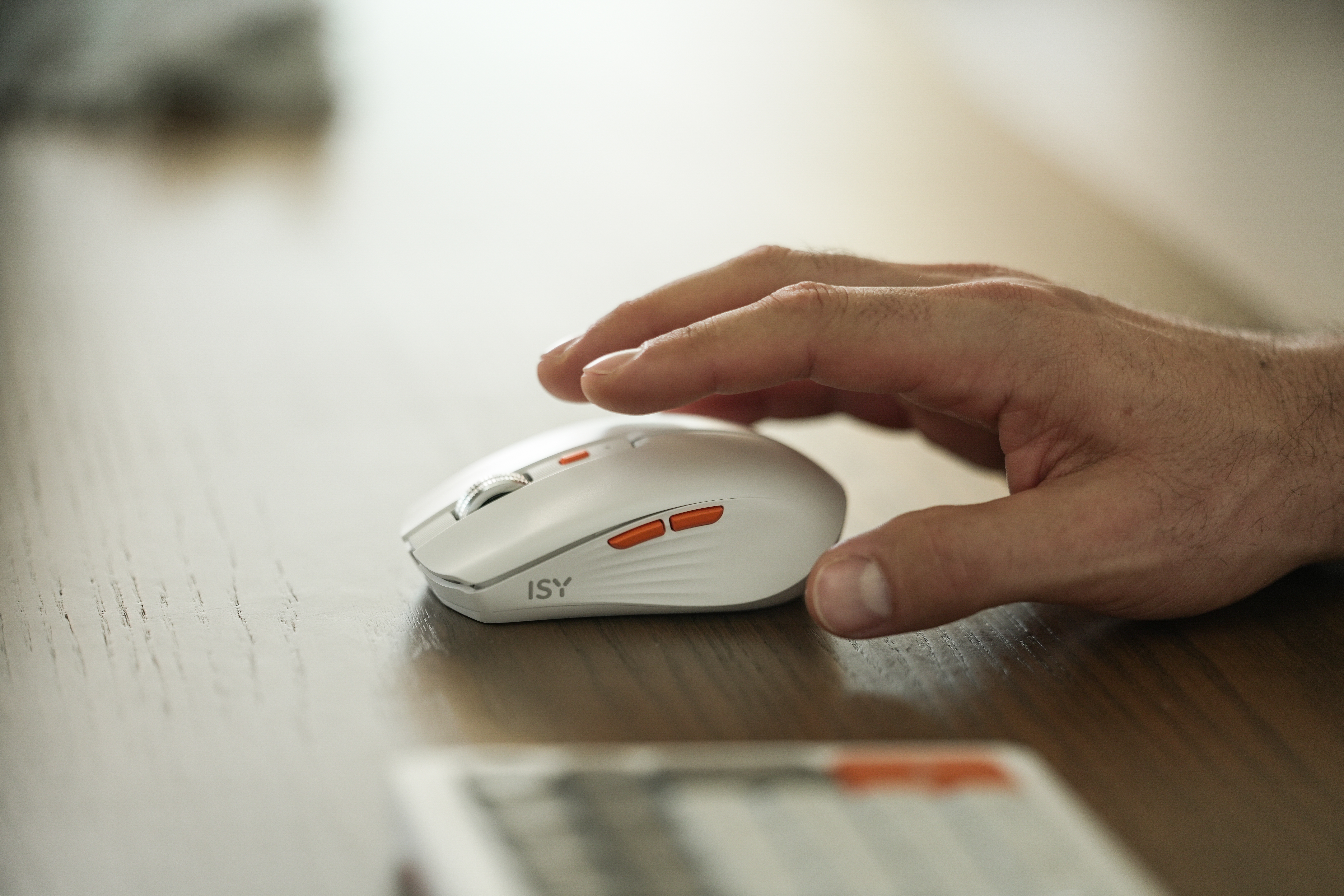 Eine Hand schwebt über einer weißen Computermaus mit orangefarbenen Akzenten. Die Maus liegt auf einer Holzoberfläche.
