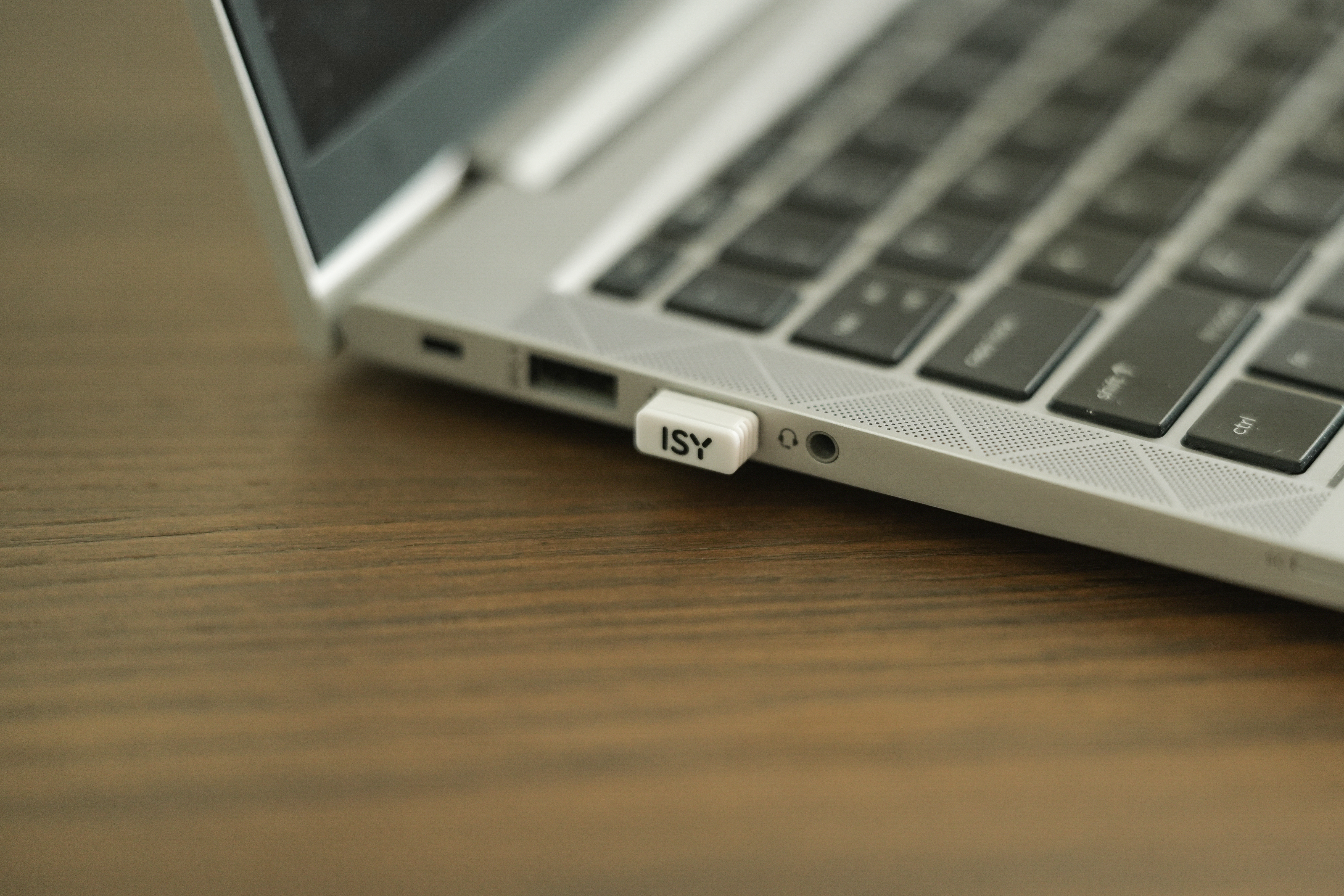 Ein Laptop mit einem weißen USB-Laufwerk mit der Aufschrift 'ISY' liegt auf einem Holztisch. Die Tastatur ist im Fokus.
