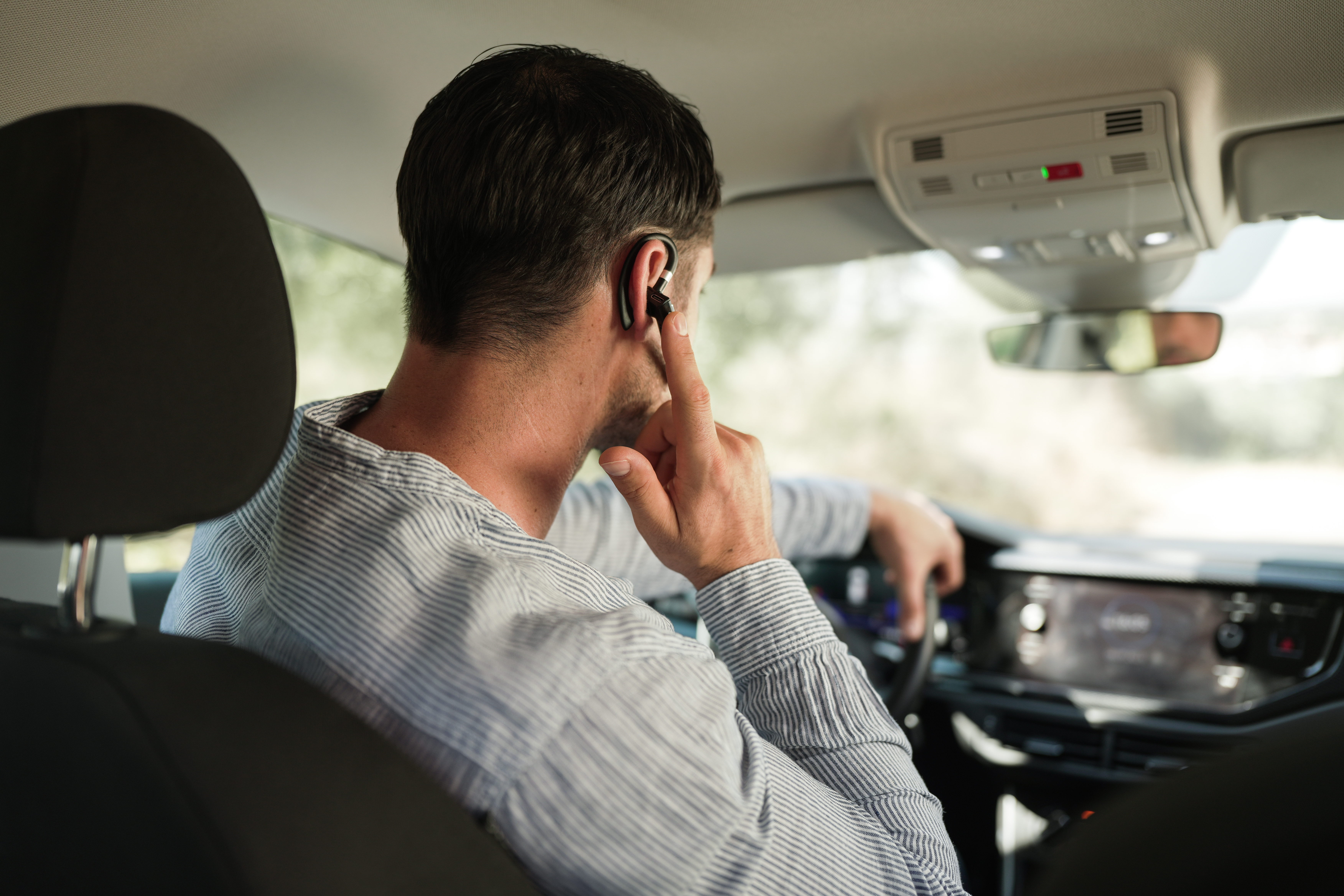 Mann im Auto, mit Headset, berührt es. Innenaufnahme. Armaturenbrett, Lenkrad und Fenster sichtbar.
