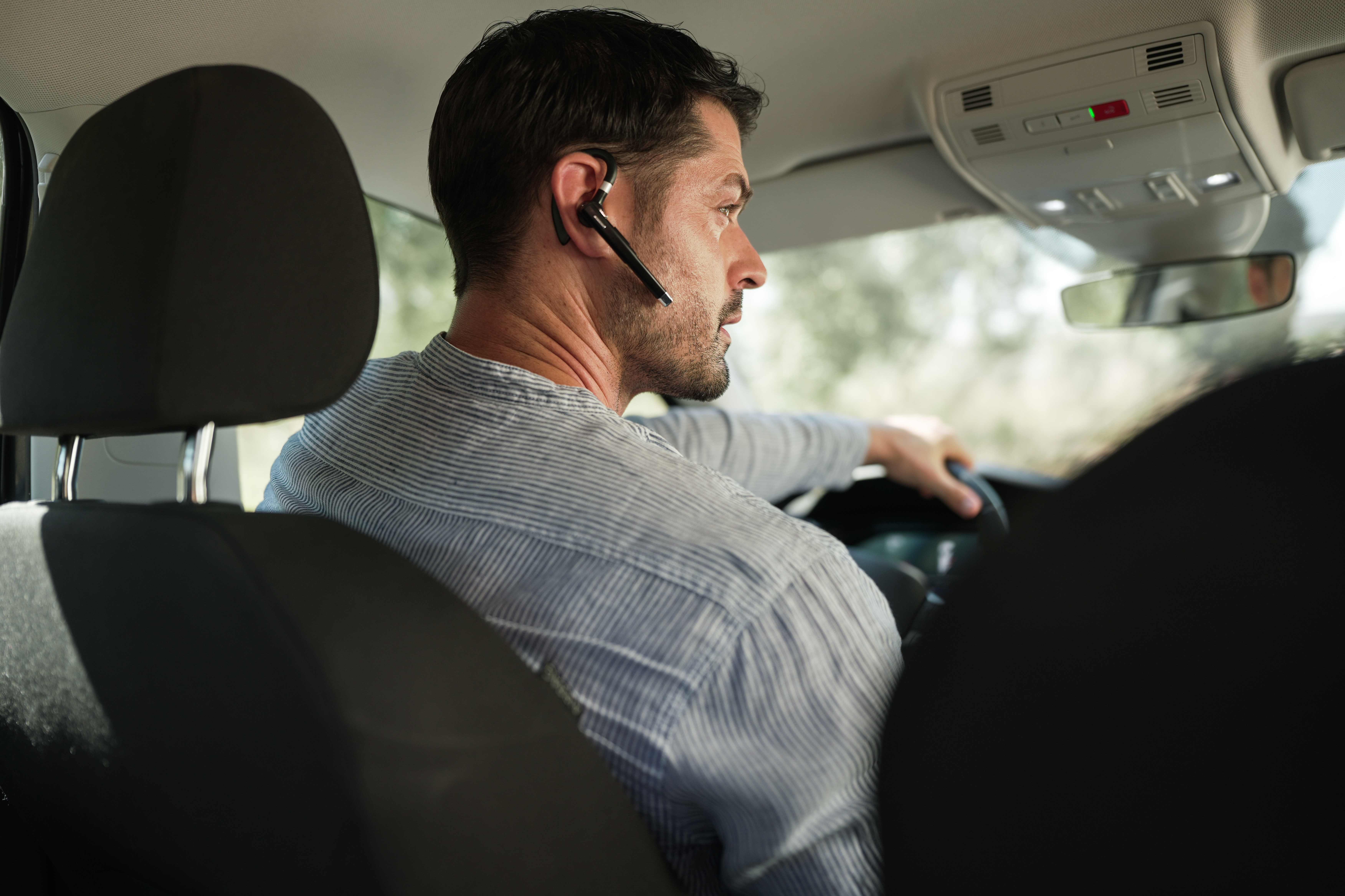 Ein Mann fährt Auto und trägt ein Headset. Er blickt nach vorne. Der Innenraum des Autos ist sichtbar.