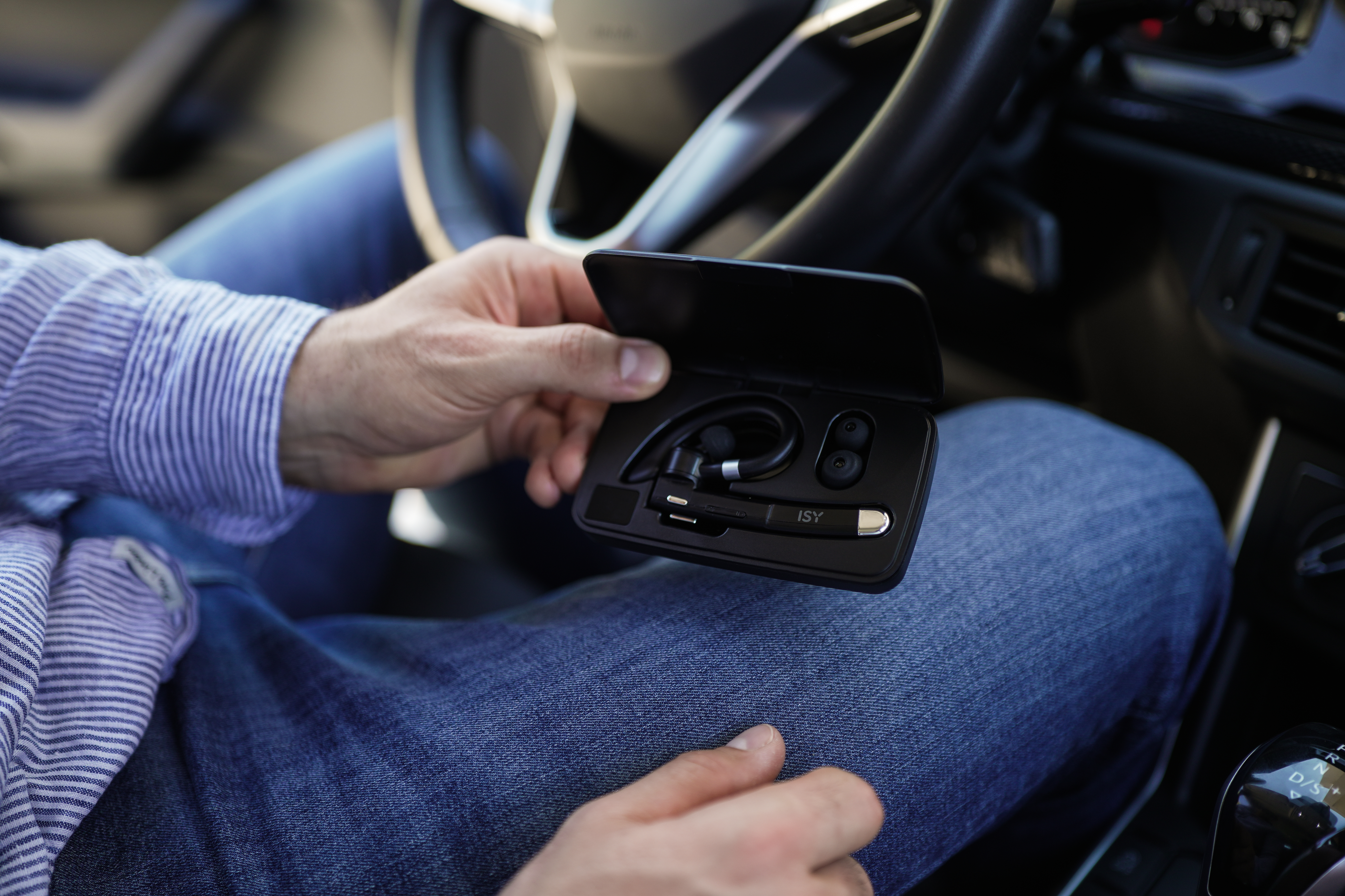 Eine Person in einem Auto hält eine schwarze ISY Bluetooth-Headset-Hülle. Die Person trägt ein blau gestreiftes Hemd und Jeans.