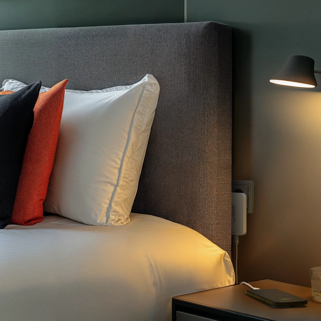 Ein Bett mit weißer Bettwäsche, Kissen, einem schwarzen und einem orangefarbenen Kissen. Eine Lampe scheint auf einen Nachttisch mit einem ladenden Telefon.