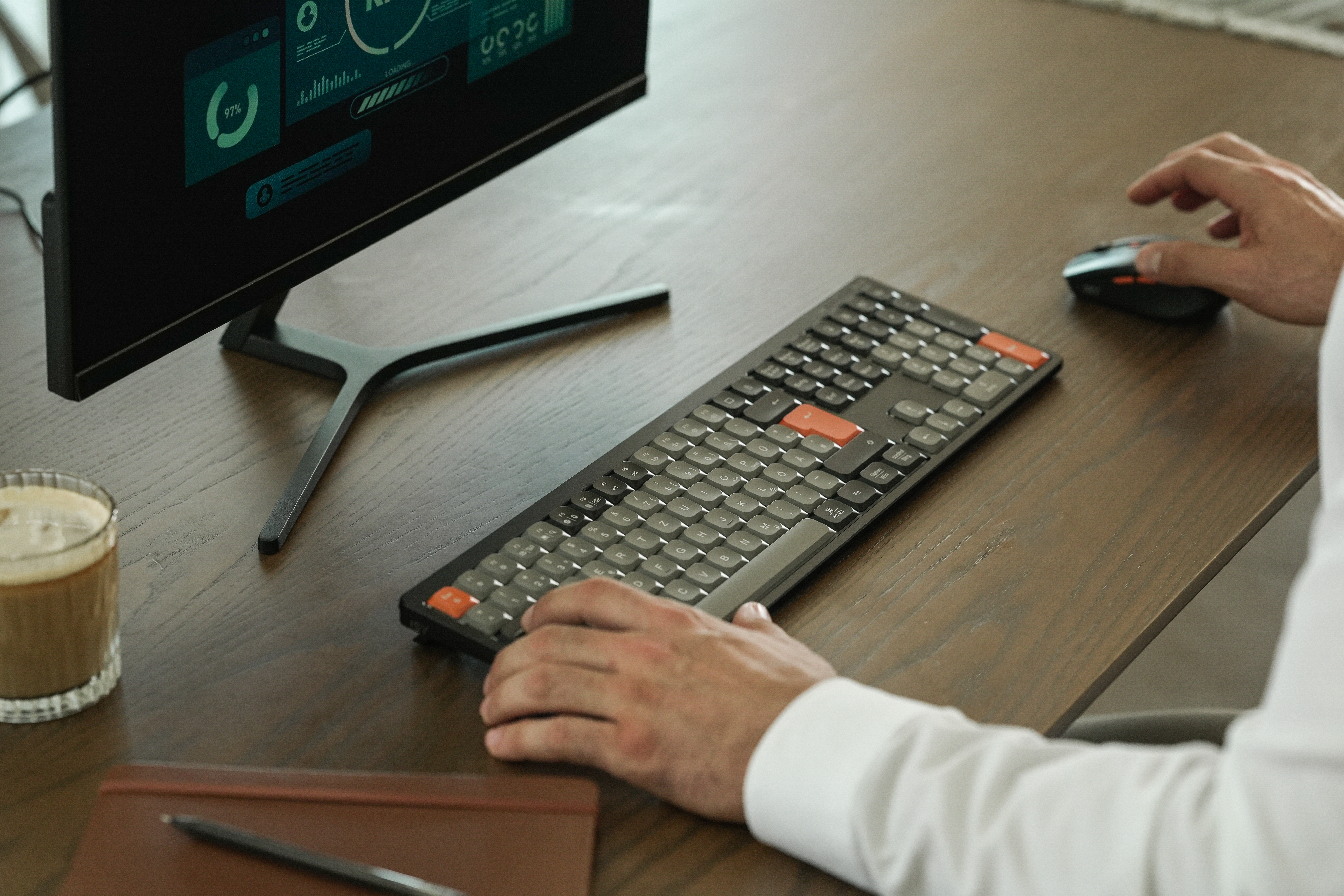 Eine Person benutzt eine Tastatur und Maus auf einem Holzschreibtisch. Ein Monitor zeigt Daten an. Kaffee und Notizblock sind vorhanden.