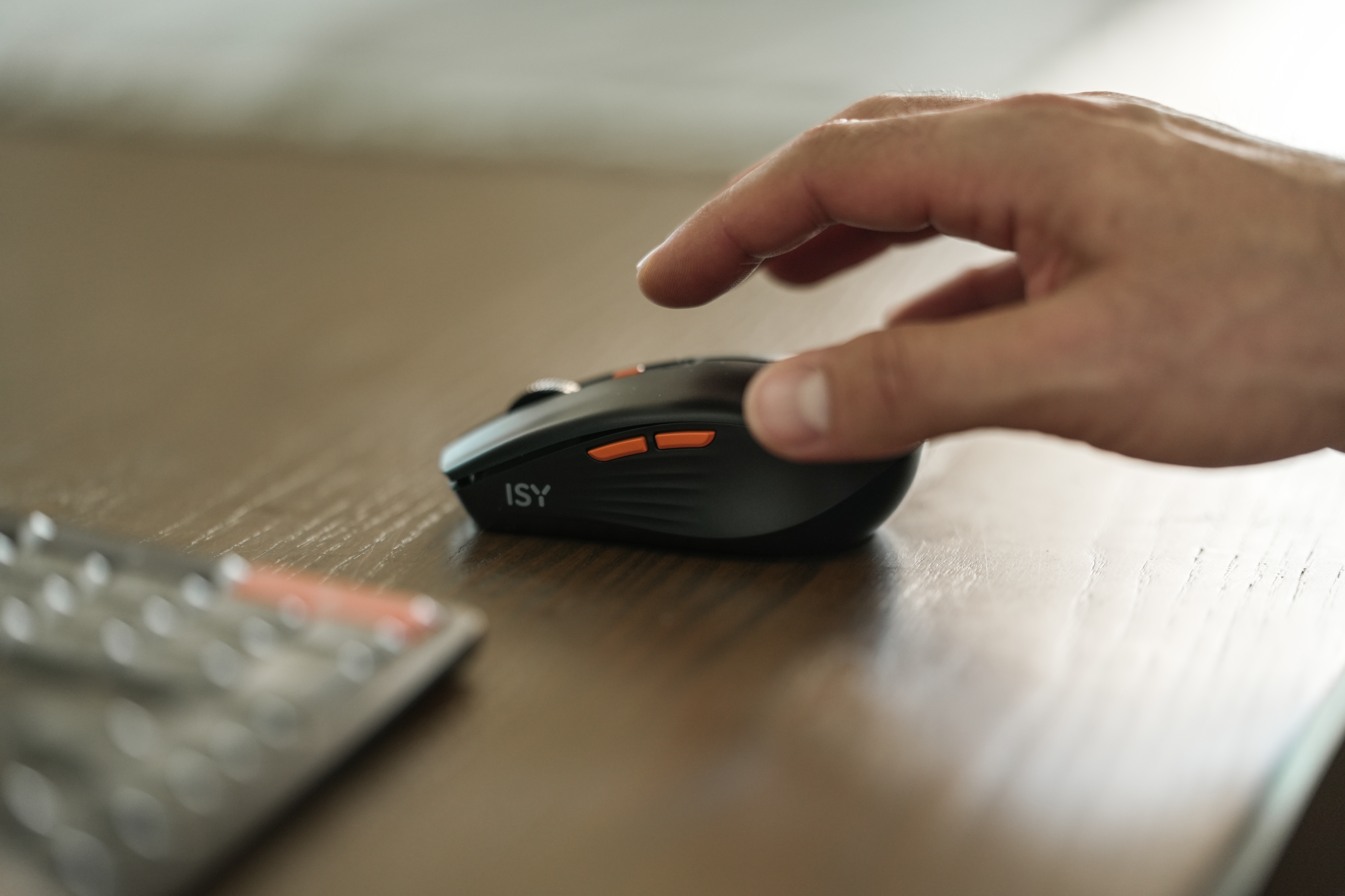 Eine Hand schwebt über einer schwarzen Computermaus mit orangefarbenen Tasten auf einem Holztisch. Eine Tastatur ist im Vordergrund.