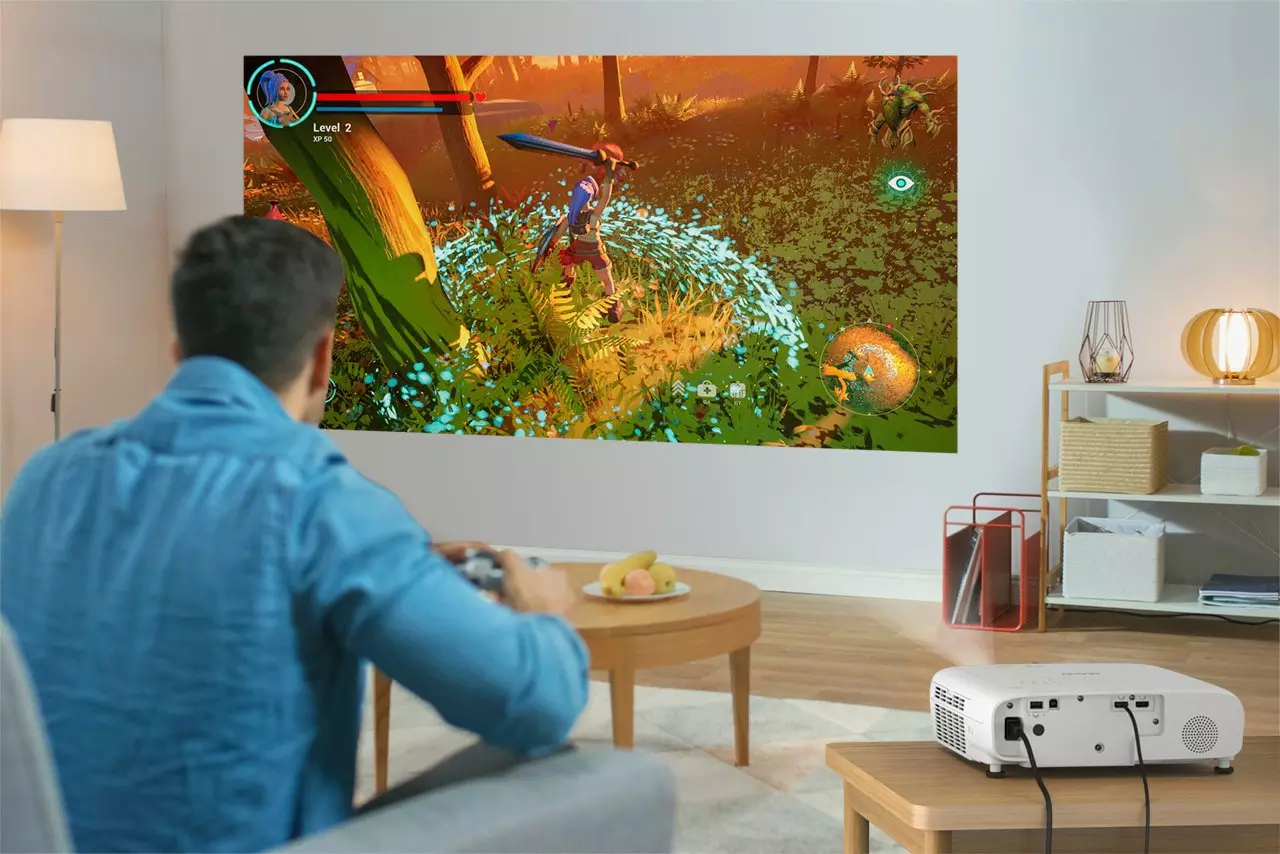 Egy férfi egy falra vetített videojátékot játszik. A játék egy kardos karaktert mutat egy erdei jelenetben. Egy projektor van az asztalon.