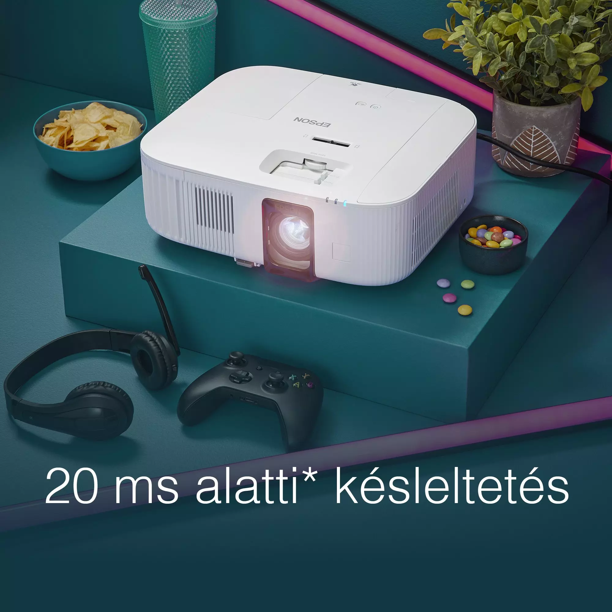 Fehér projektor zöld felületen, chips-szel, fejhallgatóval, kontrollerrel és növényekkel. Szöveg: '20 ms'.
