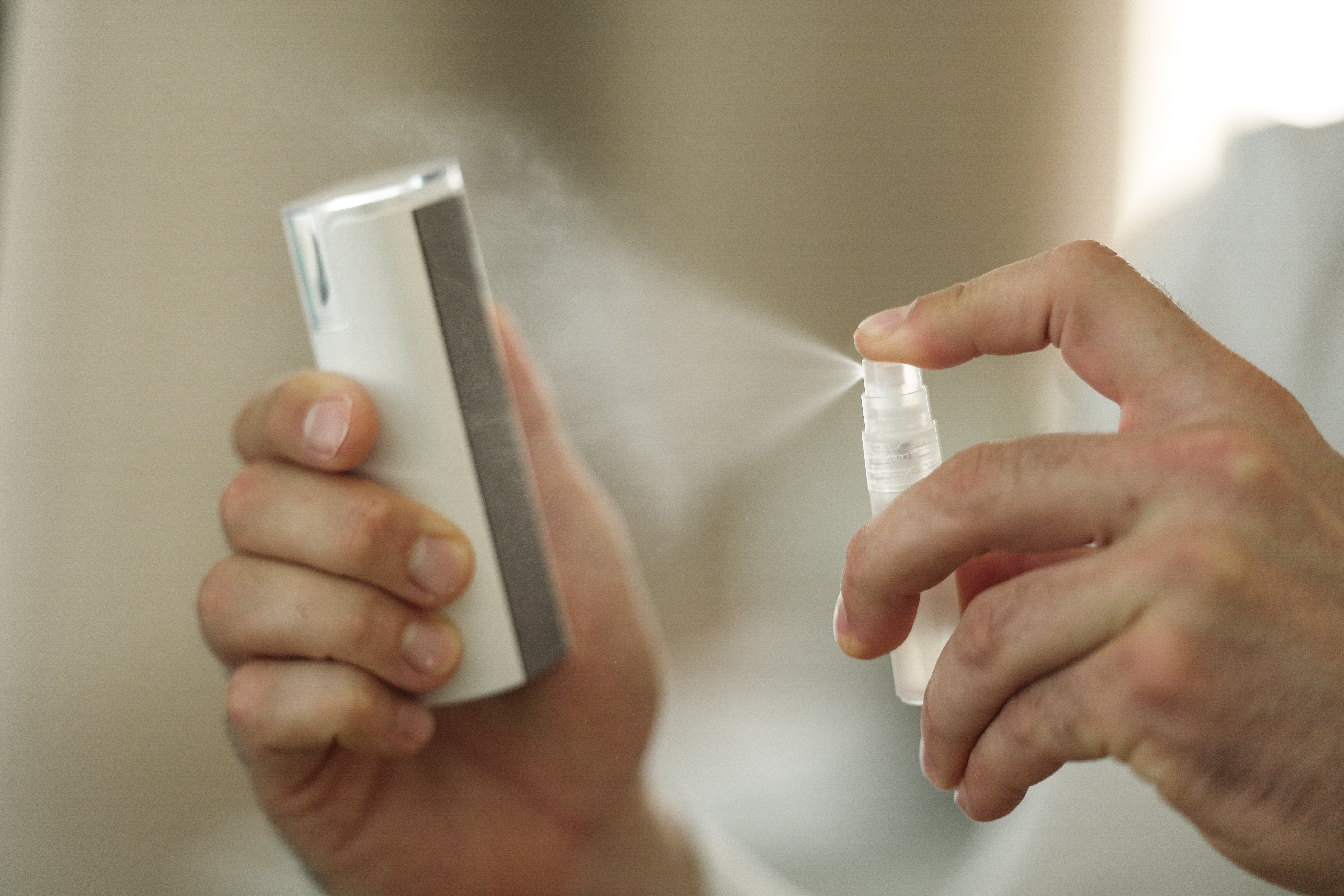 Hand sprüht Flüssigkeit aus einer kleinen Flasche, ein anderes Objekt wird in der anderen Hand gehalten. Heller Hintergrund.