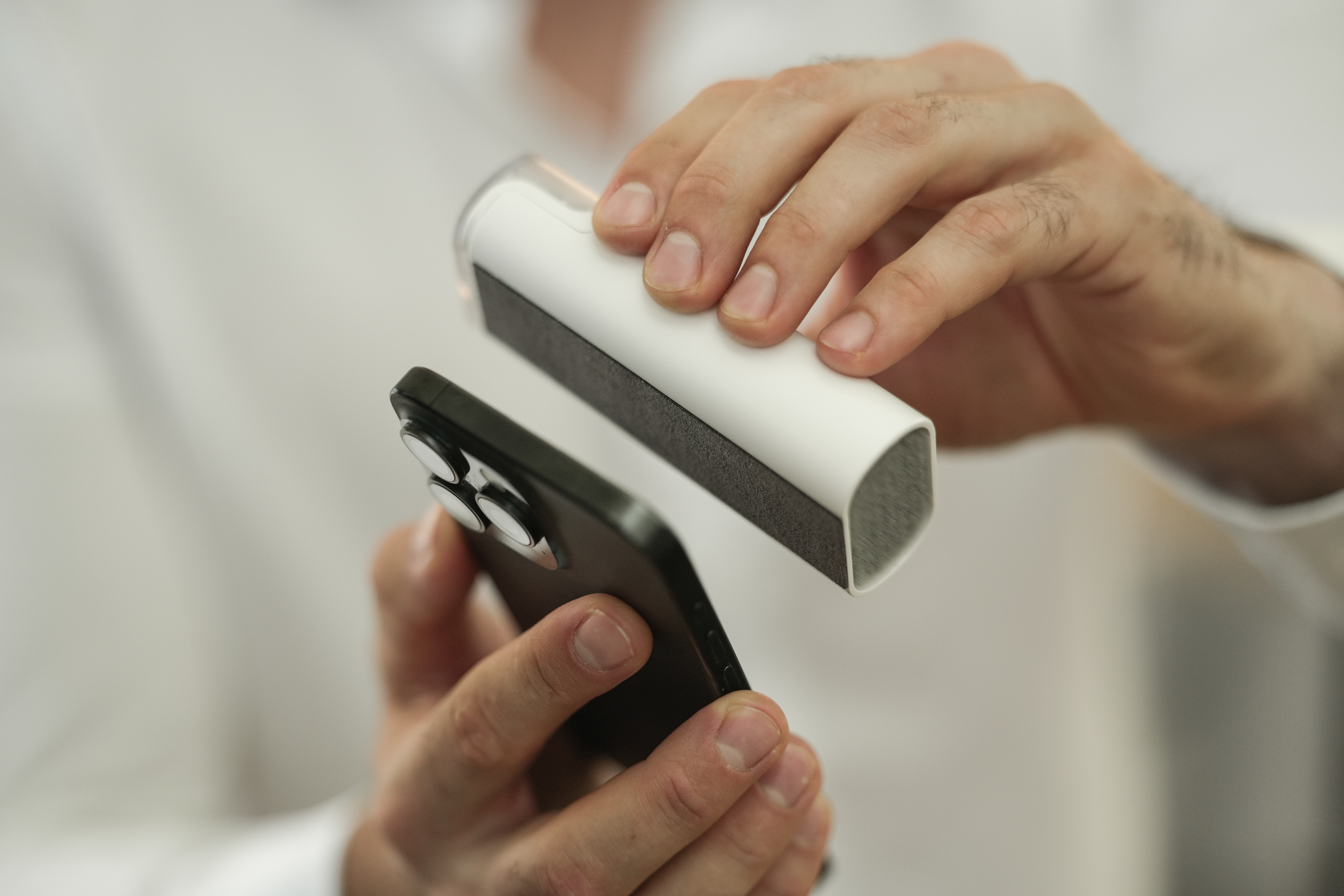Eine Person hält ein schwarzes Telefon und ein weißes Reinigungswerkzeug. Das Werkzeug wird zum Reinigen der Kameralinsen des Telefons verwendet.