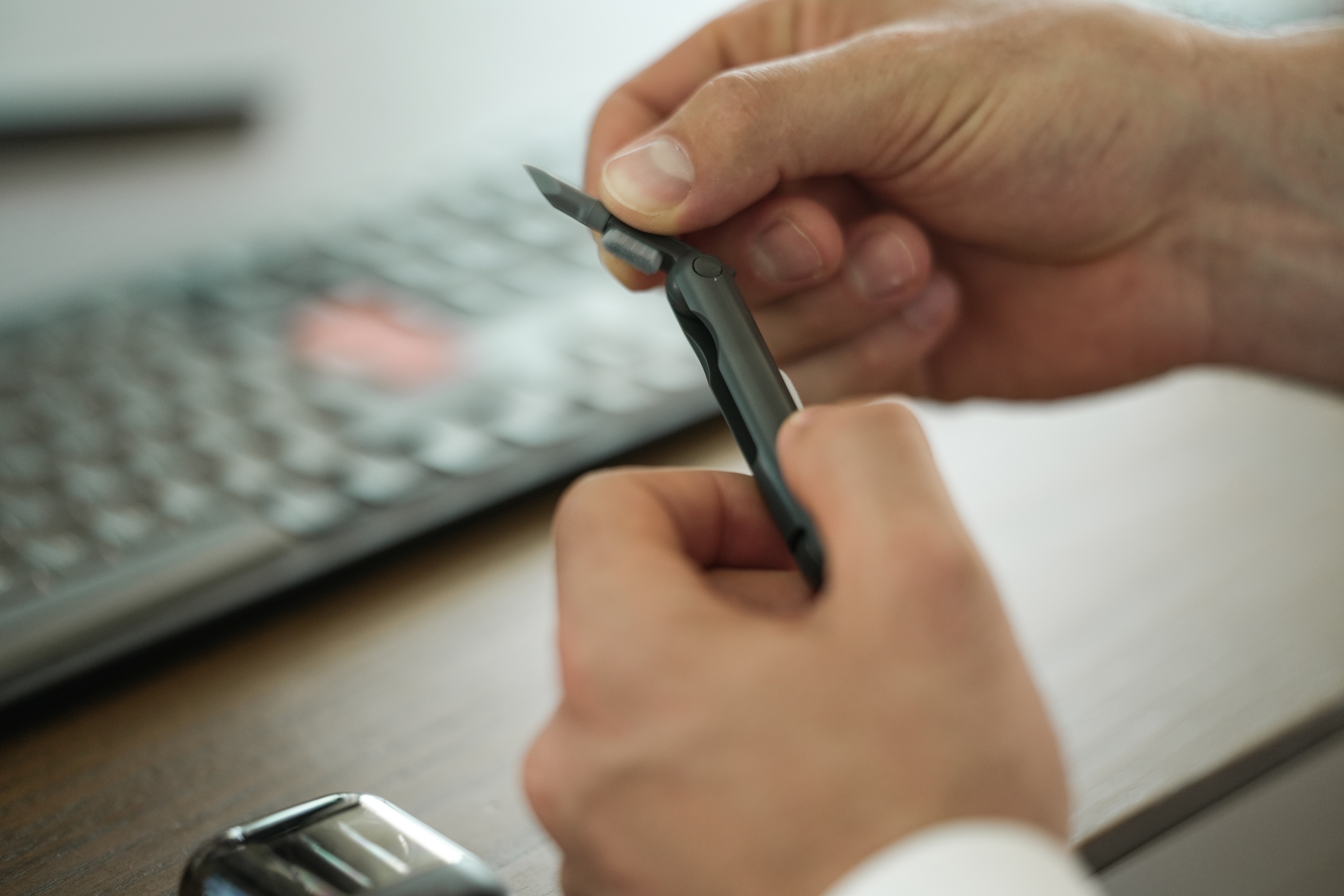 Hände halten ein kleines, dunkelgraues Messer. Im Hintergrund eine verschwommene Tastatur, darunter eine Holzoberfläche.