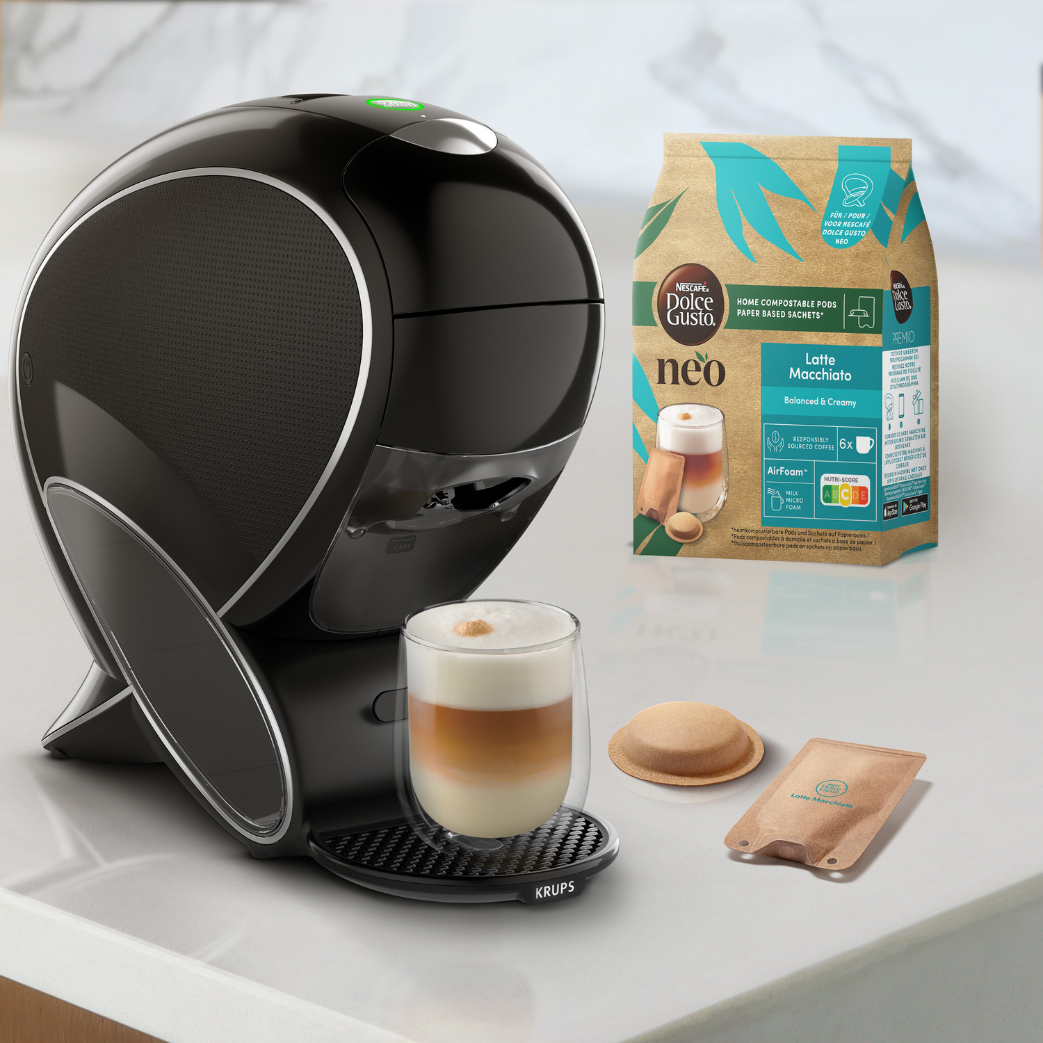 Black coffee machine with a glass of latte macchiato. A coffee pod and a packet are nearby. A coffee bag is on the right.