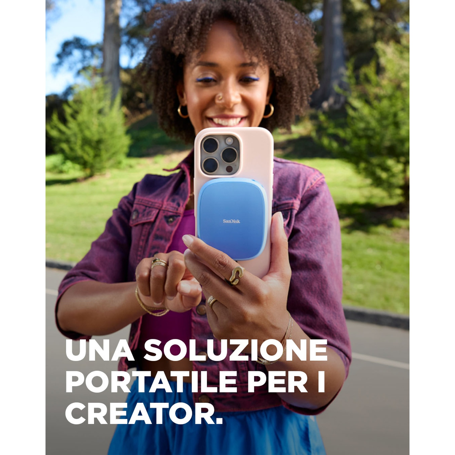 Donna con telefono e dispositivo SanDisk blu. Sorride, indossa una giacca viola. Parco verde sullo sfondo.