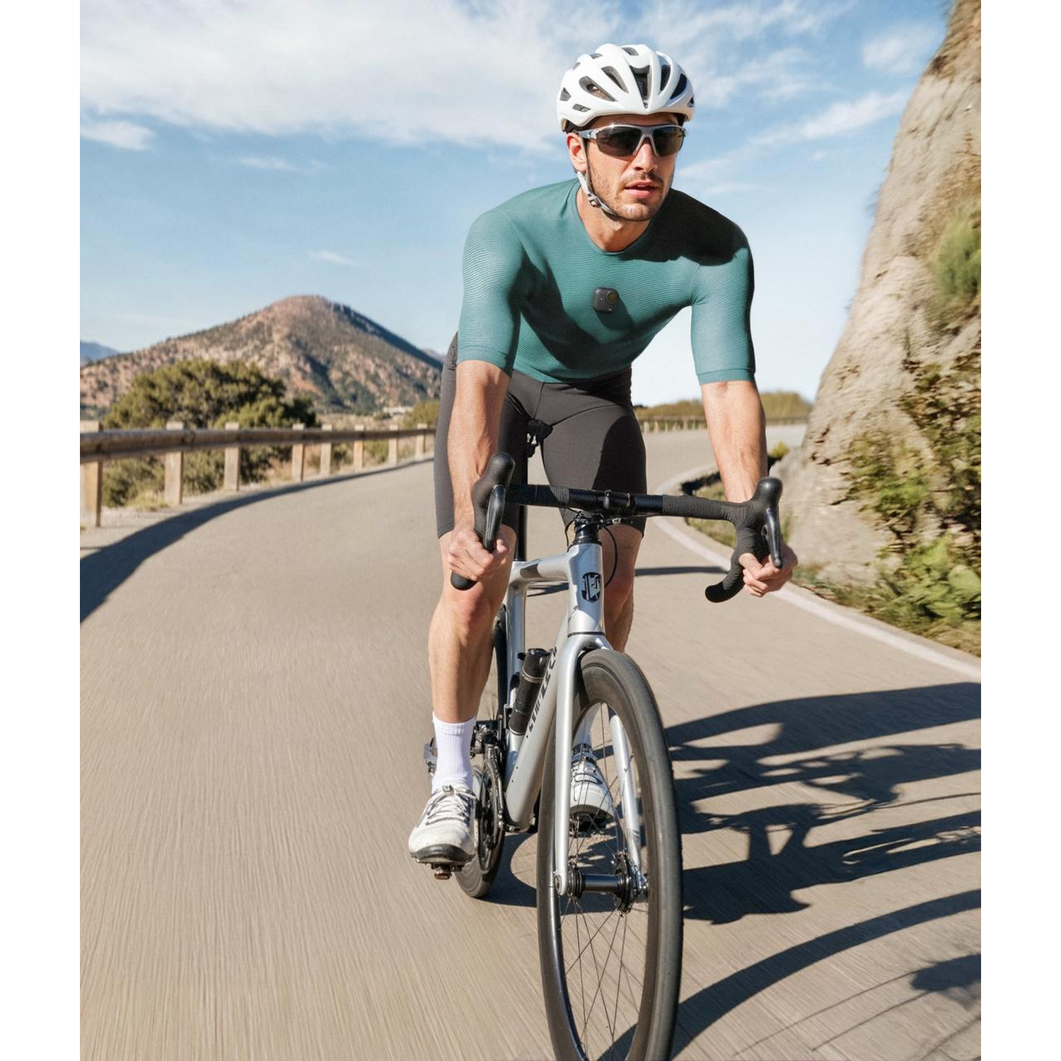 Cyclist on road, wearing helmet and sunglasses. Mountains and blue sky in background. Riding a silver bike.