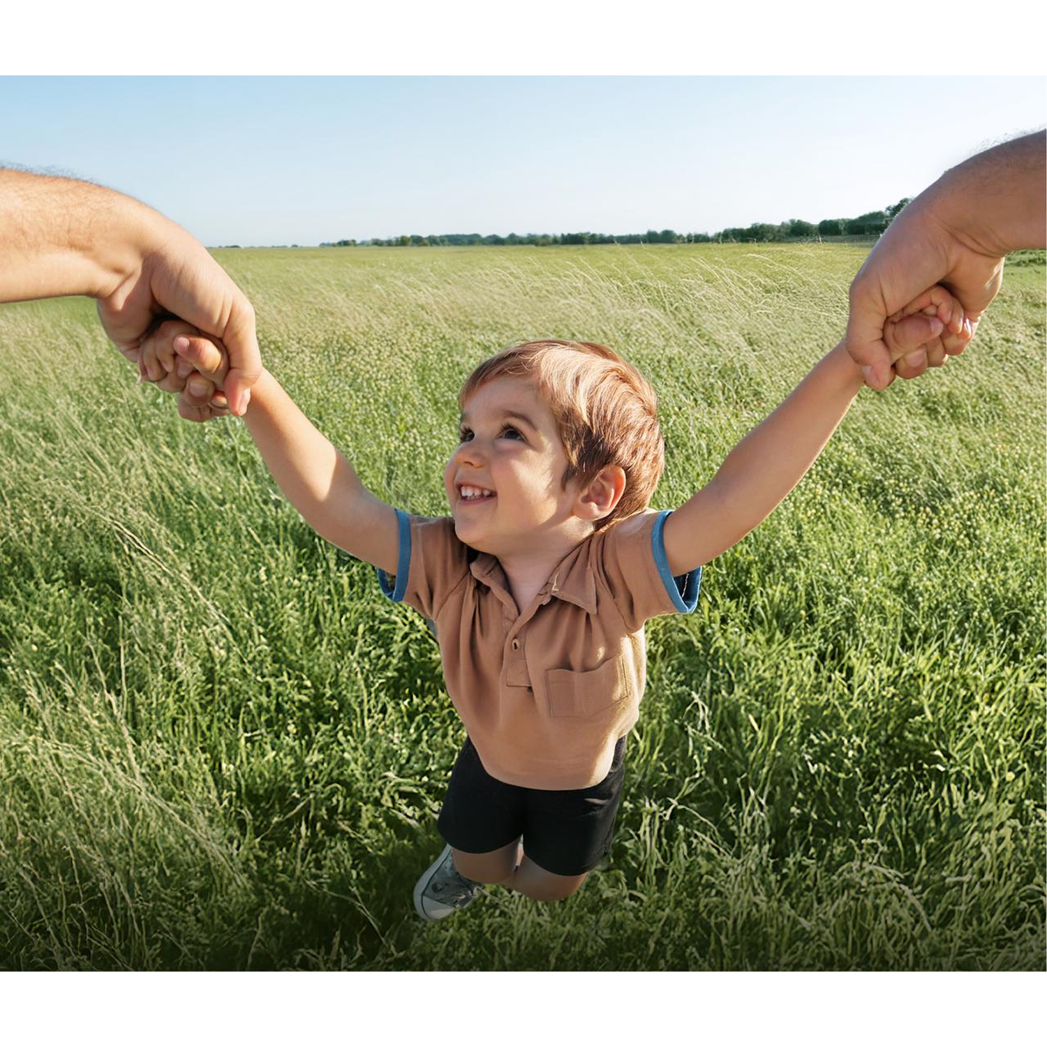 A child is held by two hands, swinging in a field. The child has brown hair and is wearing a brown shirt and black shorts.