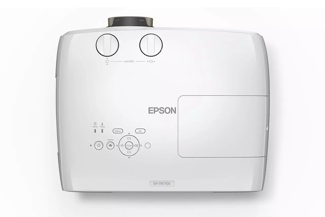 Fehér Epson EH-TW7100 projektor. Objektív eltolás tárcsák, vezérlőgombok és az Epson logója. A készülék fehér háttéren van.