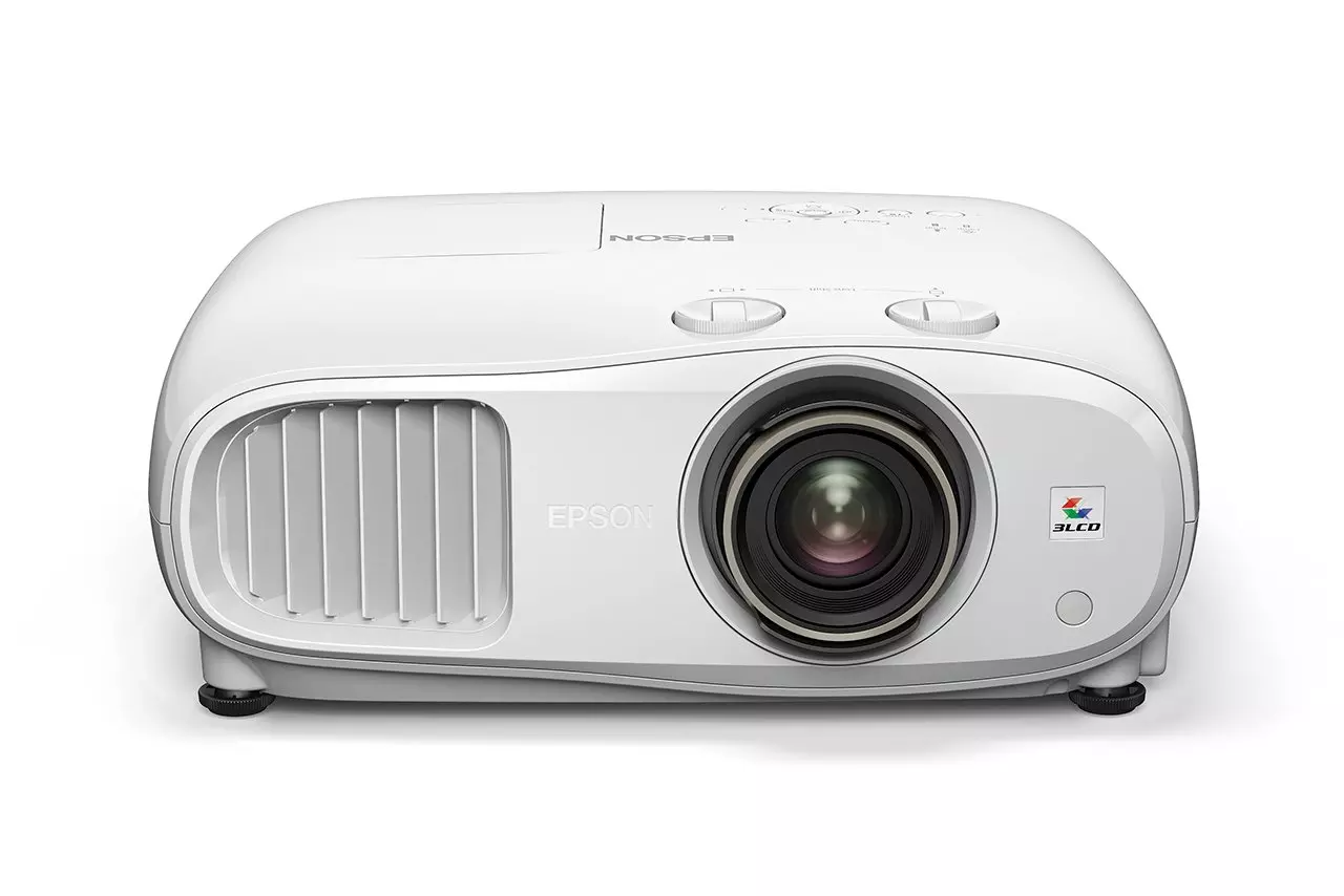 Egy fehér Epson projektor egy fehér felületen. Lencséje, szellőzőrácsai és vezérlőgombjai vannak. Az Epson logója látható.