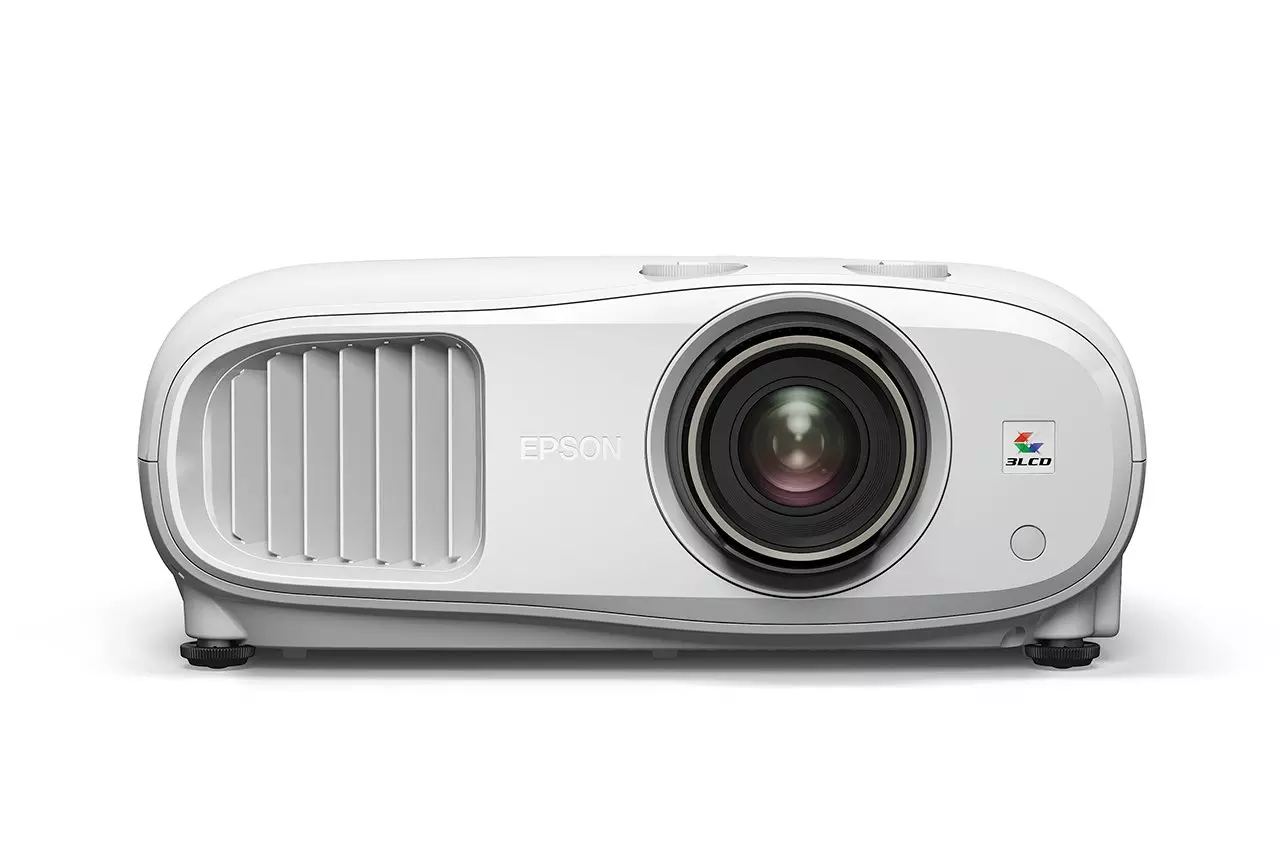 Egy fehér Epson projektor. Van rajta lencse, szellőzőnyílások és a 3LCD logó. A projektor fehér felületen van.