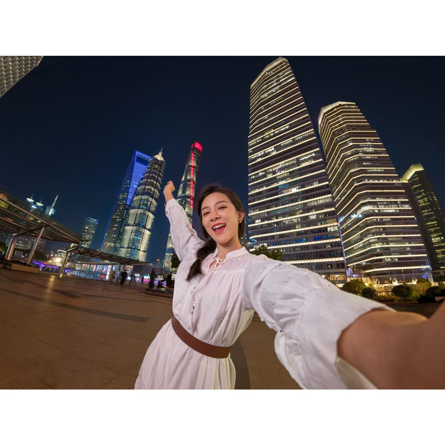 Femme prenant un selfie devant des gratte-ciel de Shanghai la nuit. Elle porte une robe blanche, une ceinture marron et sourit. Grands bâtiments en arrière-plan.