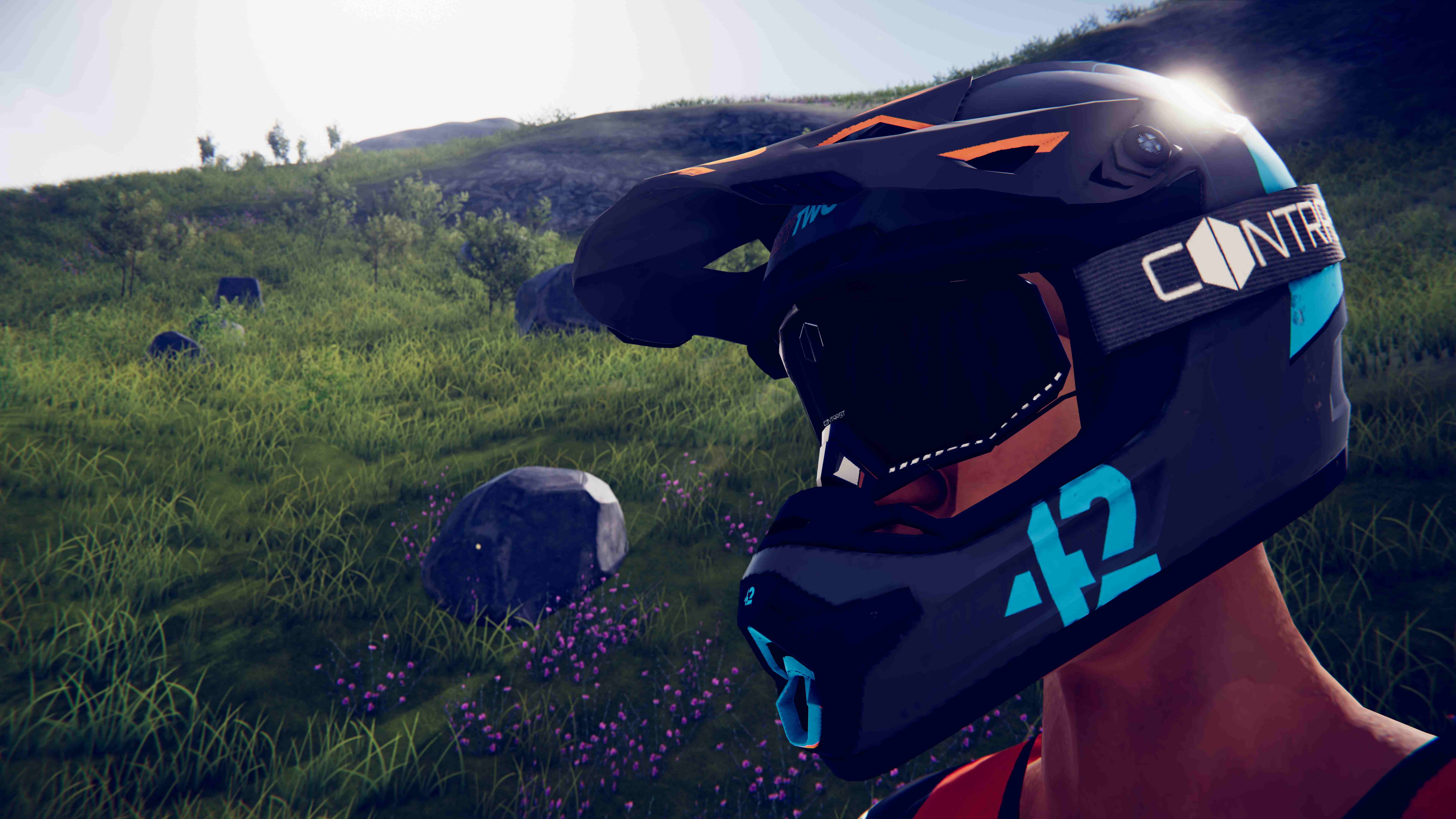 Une personne porte un casque noir avec des accents orange et turquoise. L'arrière-plan est composé d'herbe verte, de rochers et d'une colline.