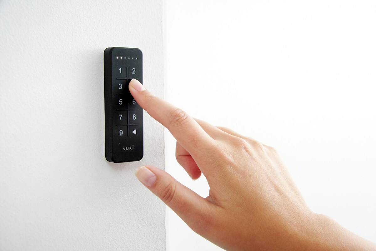 Una mano presiona el número 3 en un teclado negro montado en una pared blanca. El teclado tiene los números 1-9 y la marca Nuki.