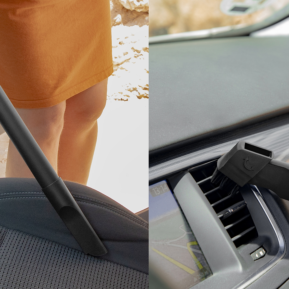 Una persona aspirando un asiento de coche. La aspiradora es negra. El salpicadero es visible y las rejillas de ventilación se limpian con un cepillo.