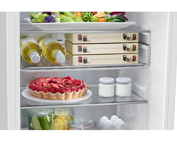 Innenraum eines Kühlschranks mit Salat, Wein, Schokoladenbechern, Erdbeerkuchen, Joghurt, Gurken, Zitronen und Eiern.