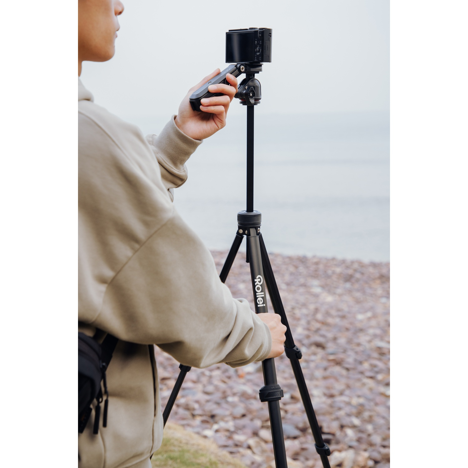 A person with a camera on a tripod near a rocky shore. The person is holding a remote control. The sky is overcast.