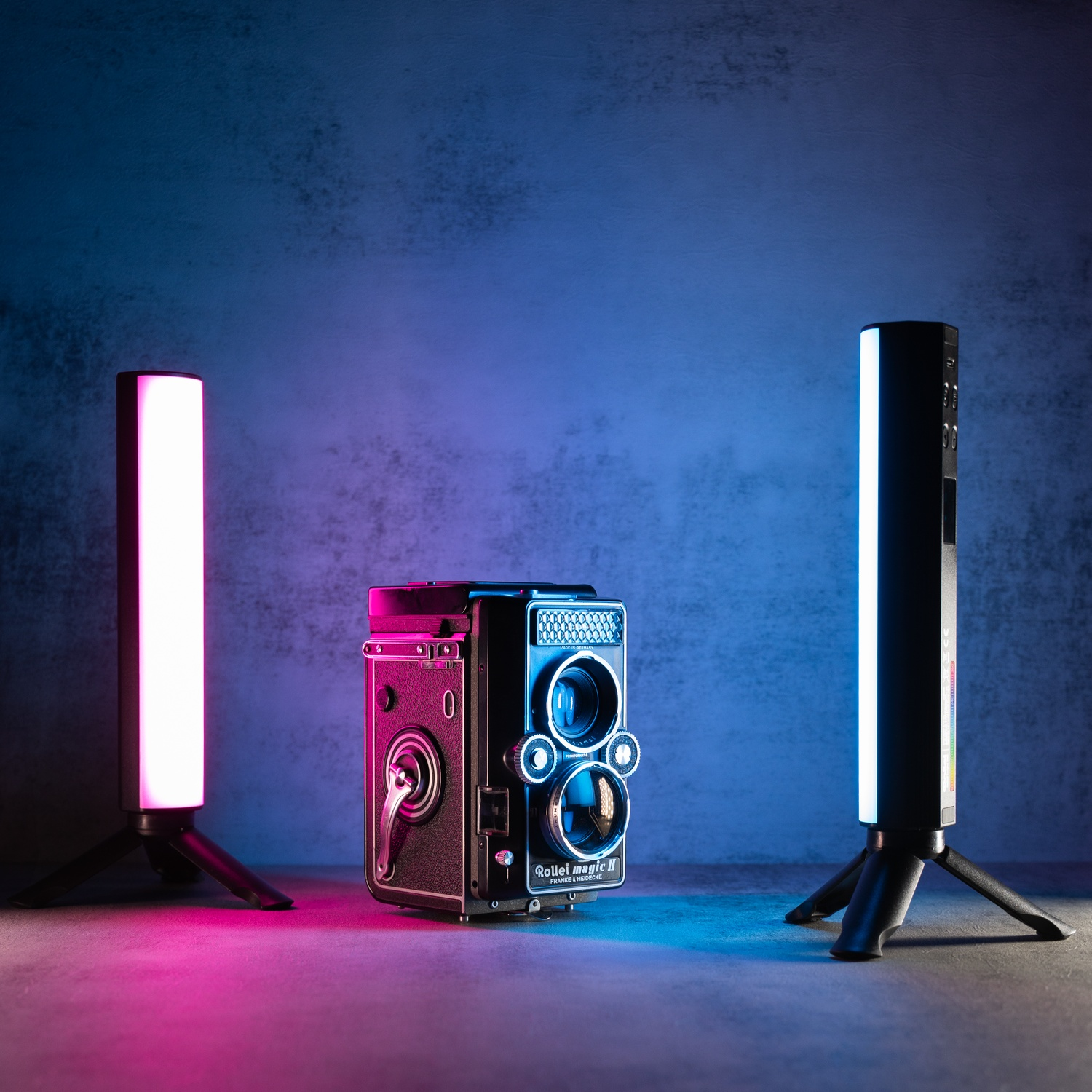 Un appareil photo vintage est flanqué de deux barres lumineuses éclairées sur des trépieds. Le fond est un mur gris texturé avec un éclairage bleu et rose.