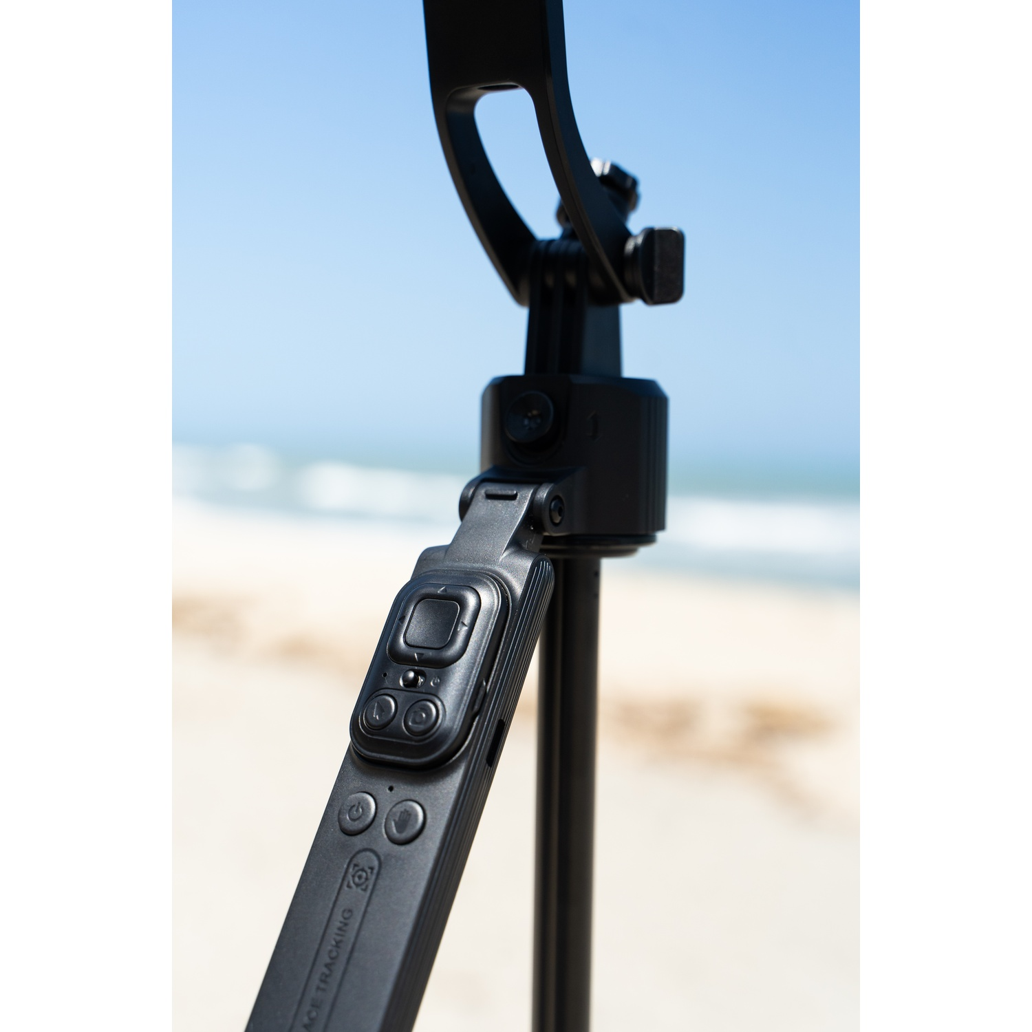 Een zwarte camera afstandsbediening met knoppen is bevestigd aan een zwart statief. De achtergrond is een strand en blauwe lucht.