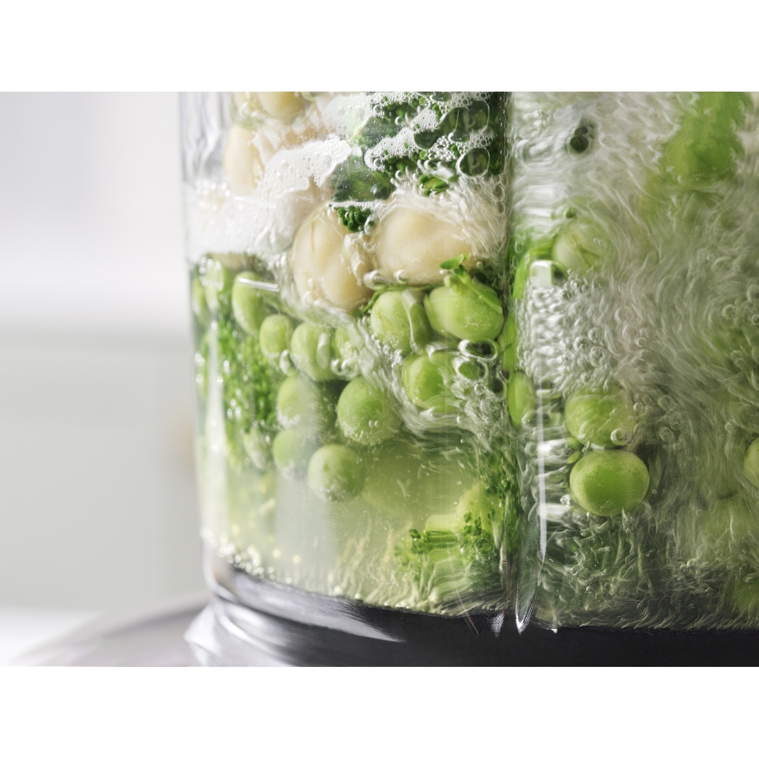 Primo piano di un frullatore pieno di piselli, ceci ed erbe in acqua. Le bolle si formano intorno agli ingredienti.