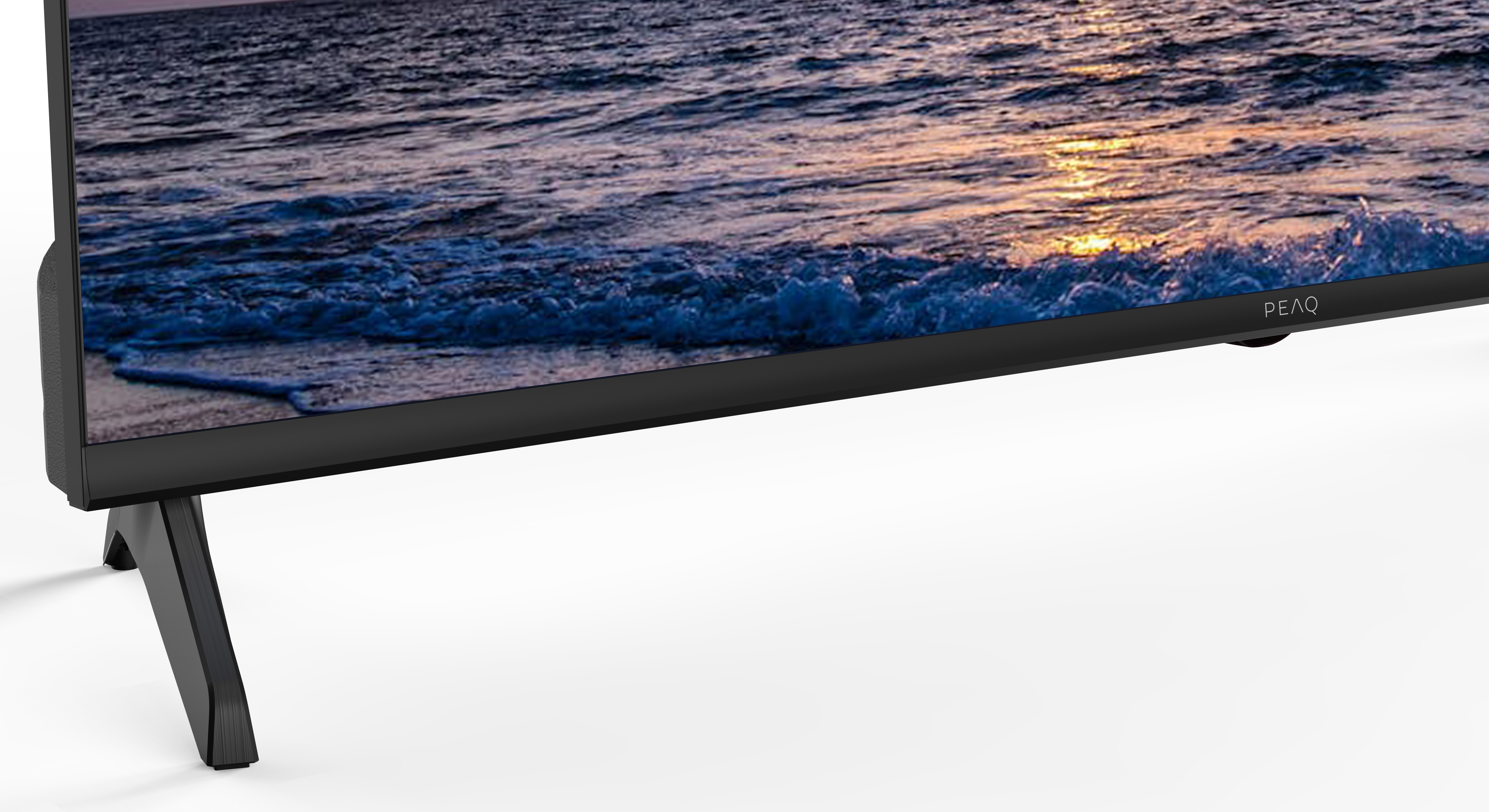 Czarny telewizor wyświetla fale oceanu o zachodzie słońca. Telewizor stoi na dwóch kątowych czarnych nogach. Widoczna jest nazwa marki PEAQ.