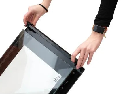 Ręce trzymają szklane drzwi piekarnika z czarną ramą. Jedna ręka nosi zegarek. Drzwi mają symbol ostrzegawczy.