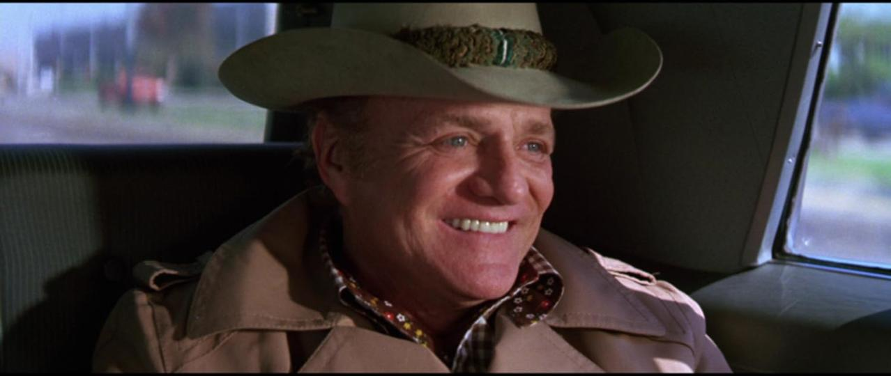 Ein lächelnder Mann mit Cowboyhut und Trenchcoat sitzt in einem Auto. Im Hintergrund sind Häuser und Bäume zu sehen.