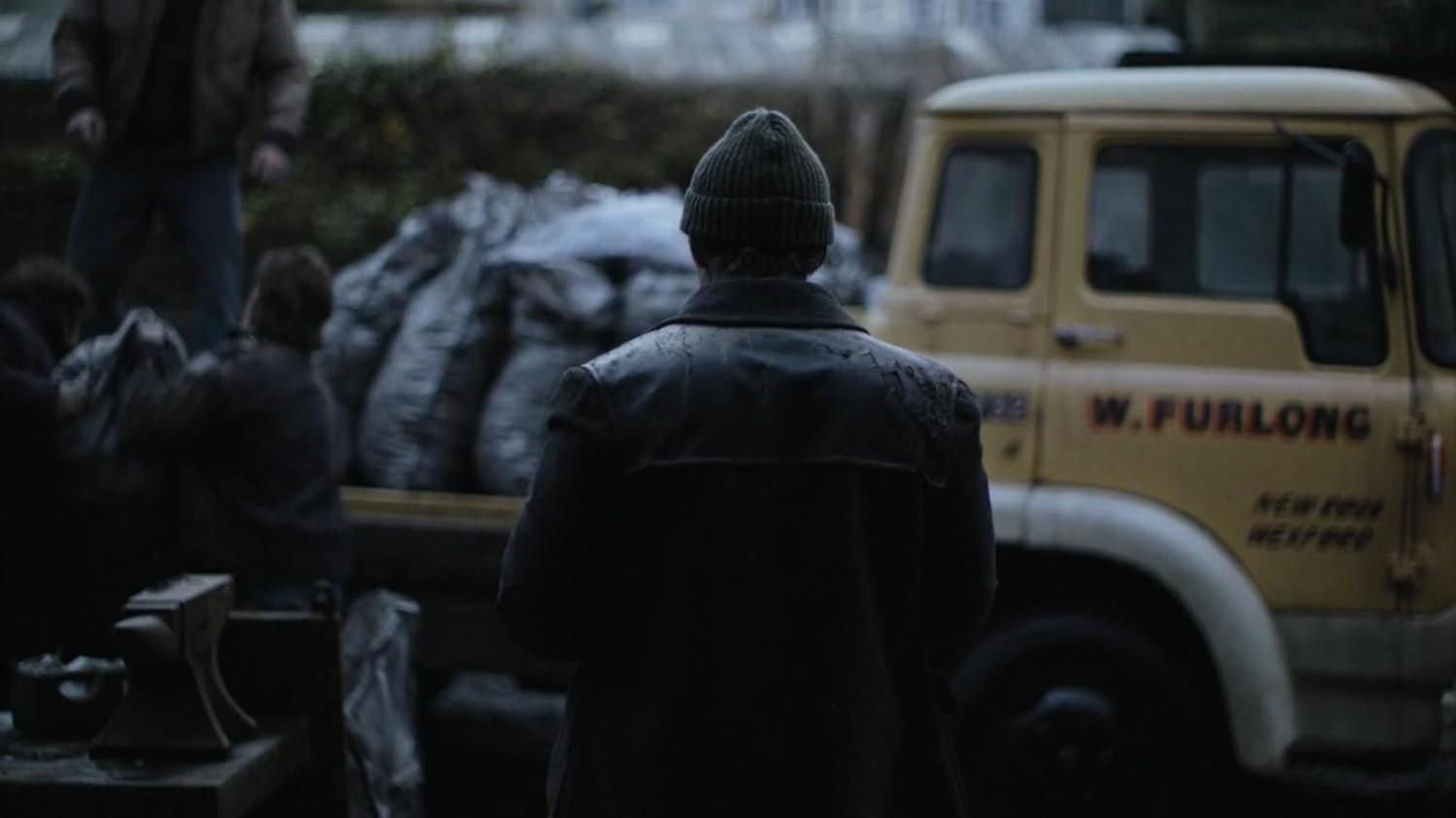 Ein Mann in Mütze und Jacke steht in der Nähe eines gelben Lastwagens mit dem Namen W. Furlong. Menschen verladen Säcke im Hintergrund.
