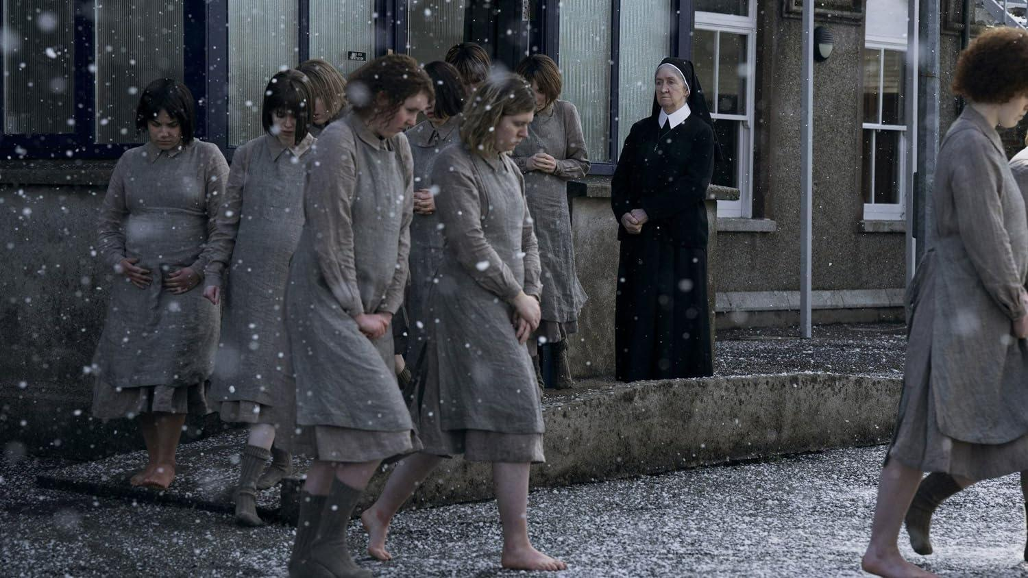 Frauen in grauen Kleidern gehen barfuß im Schnee. Eine Nonne beobachtet. Gebäude und fallender Schnee im Hintergrund.
