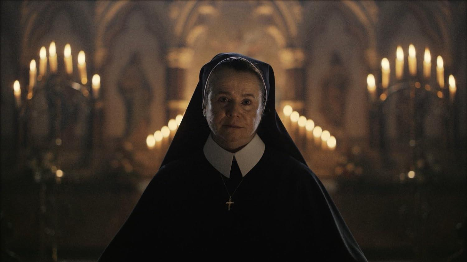 Eine Nonne in schwarzem Habit, weißem Kragen und Kreuzkette steht in einer Kirche. Auf beiden Seiten sind Kerzen im Hintergrund.