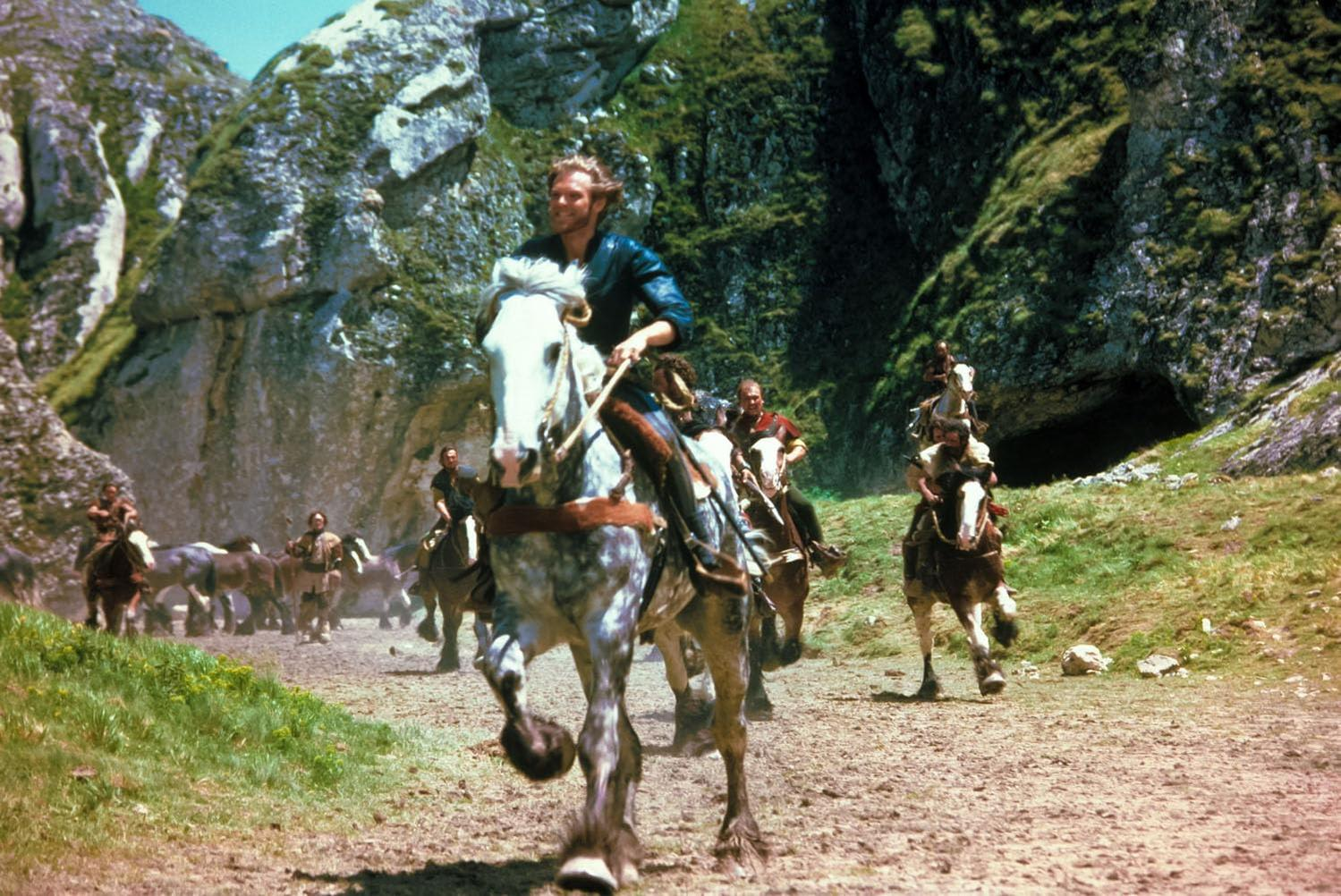 Männer auf Pferden reiten durch einen Bergpass. Der Anführer sitzt auf einem weißen Pferd. Die Szene ist sonnig.