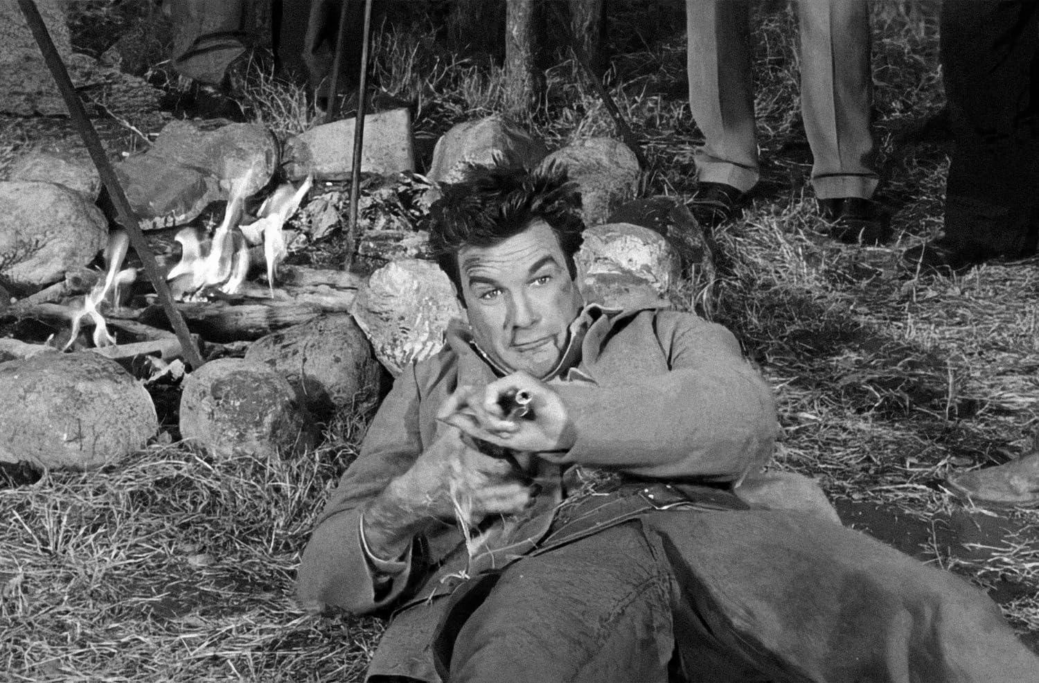 Ein Mann, der eine Waffe anvisiert, während er in der Nähe eines Lagerfeuers liegt. Felsen und Gras sind im Vordergrund.