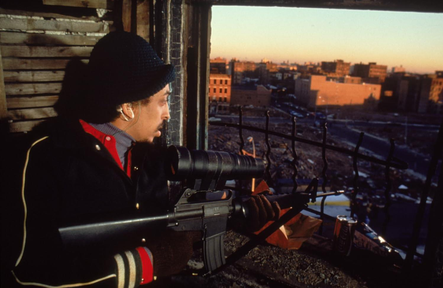 Ein Mann zielt mit einem Gewehr aus einem Fenster. Gebäude und Himmel im Hintergrund. Er trägt einen schwarzen Hut und Mantel und wirkt konzentriert.