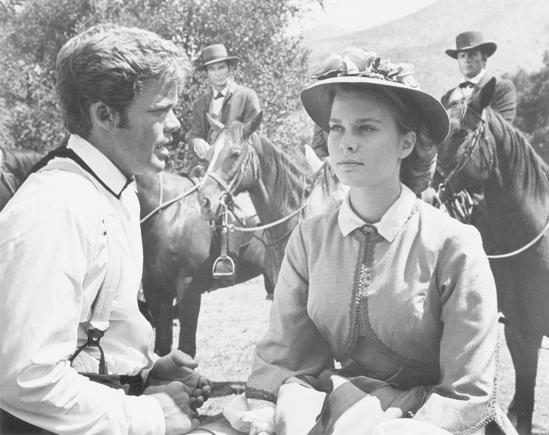 Ein Mann und eine Frau in Vintage-Kleidung. Mehrere Personen auf Pferden im Hintergrund. Schwarz-Weiß-Bild.