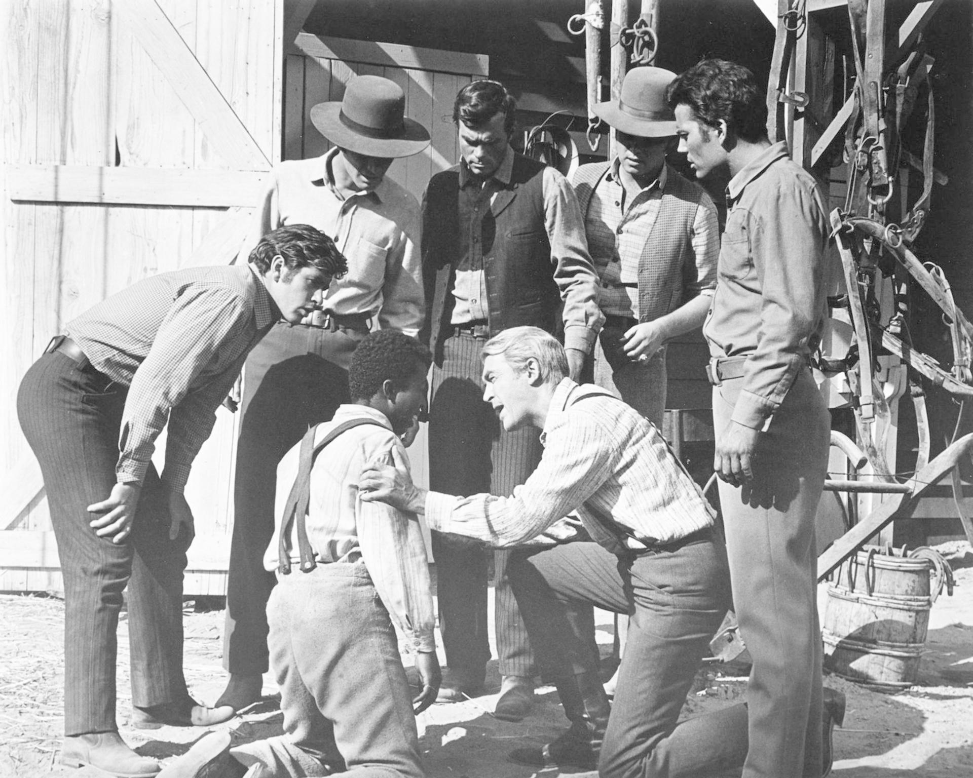 Eine Gruppe von Männern in Westernkleidung umringt zwei kniende Männer. Ein Mann tröstet den anderen. Holzstrukturen und ein Geschirr sind im Hintergrund zu sehen.
